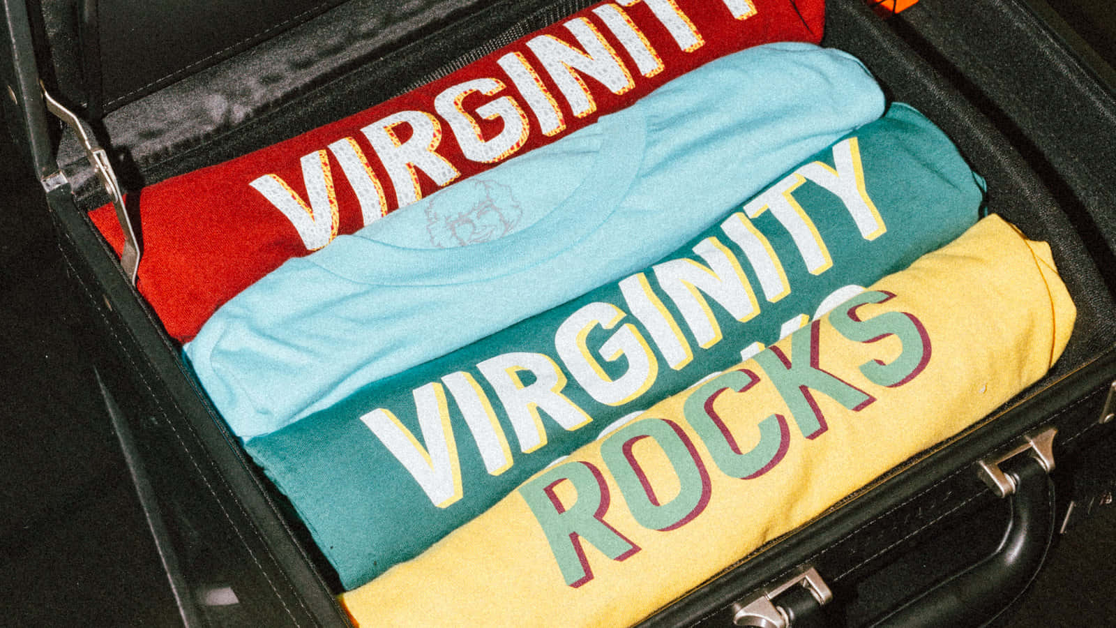  壁紙 — 整齊折疊的彩色T恤，上面印有粗體 'VIRGINITY' 和 'VIRGINITY ROCKS' 字樣，放在一個打開的黑色行李箱內。