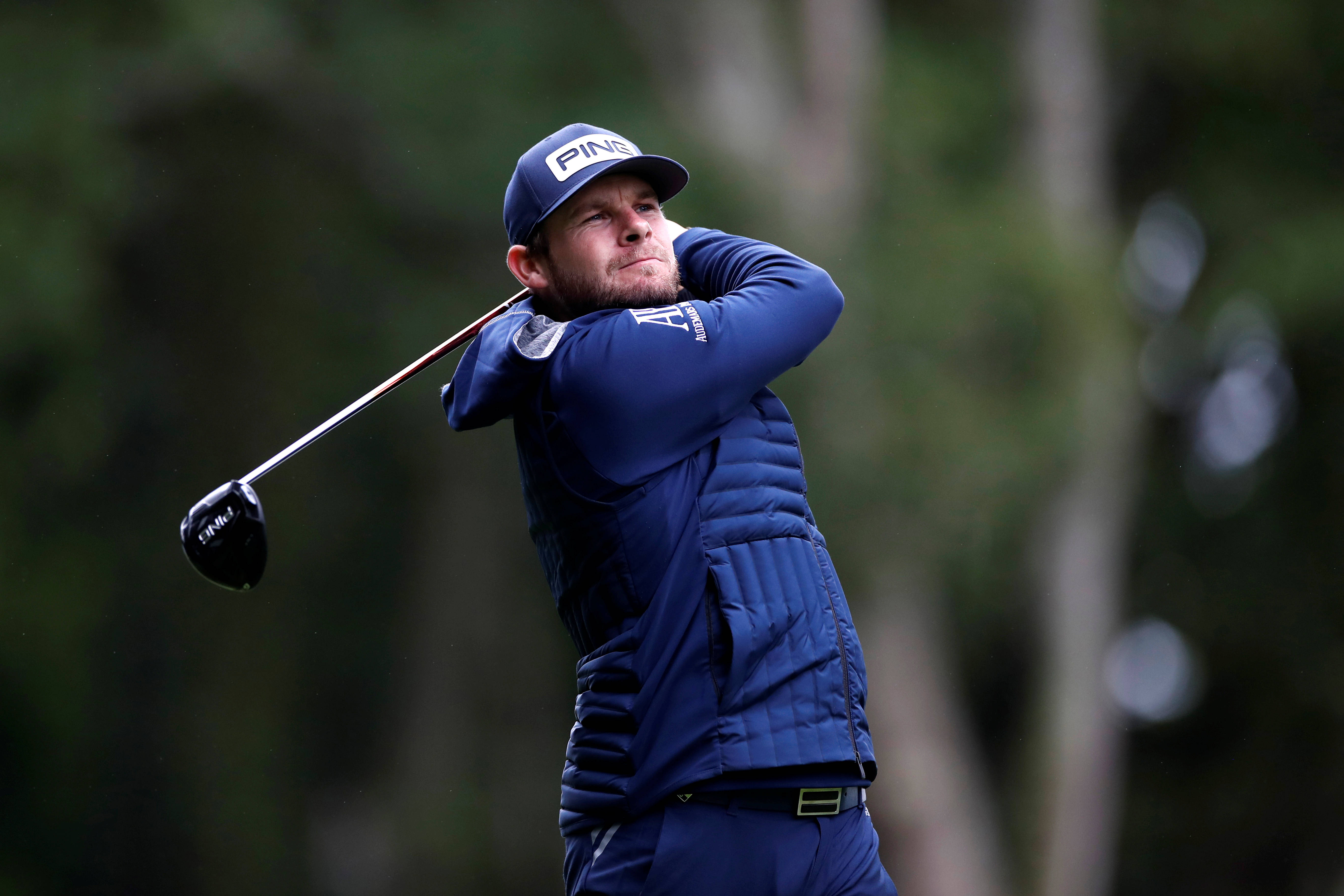 Tyrrell Hatton 桌布 壁紙 — 一位專注的高爾夫球手身穿藍色服裝，在模糊的森林背景下，以強烈的專注力揮桿。