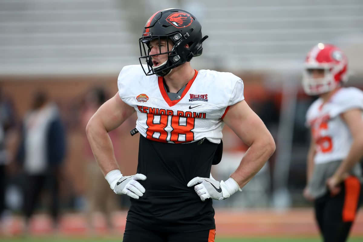 壁紙 — 一名專注的美式足球員，身穿白色和橙色的「Senior Bowl」球衣，雙手叉腰站在球場上，背景是體育館看台。