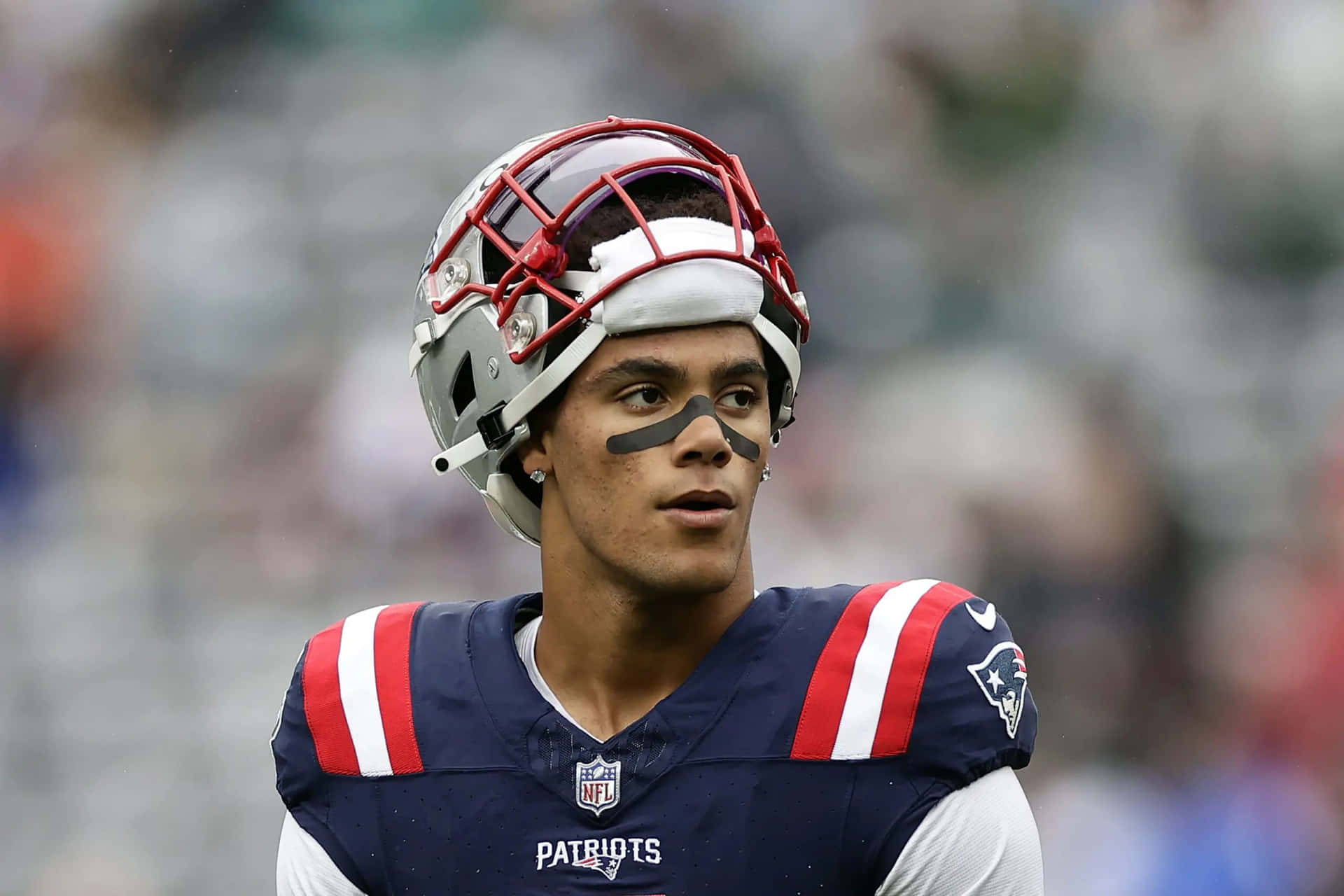  壁紙 — 一位專注的NFL美式足球員身穿愛國者隊制服，戴紅色頭盔和黑色眼影，在日間比賽中凝視。
