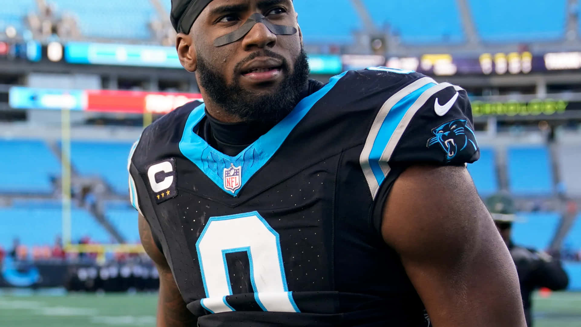  壁紙 — 一名專注的NFL球員身穿黑藍色卡羅萊納黑豹隊零號球衣，在明亮的燈光下站在球場上。