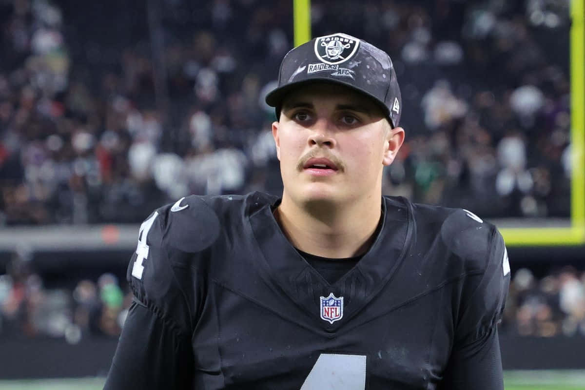  壁紙 — 一位專注的NFL四分衛身穿黑色Raiders球衣和帽子，在球場燈光下站立，背景是模糊的球迷。