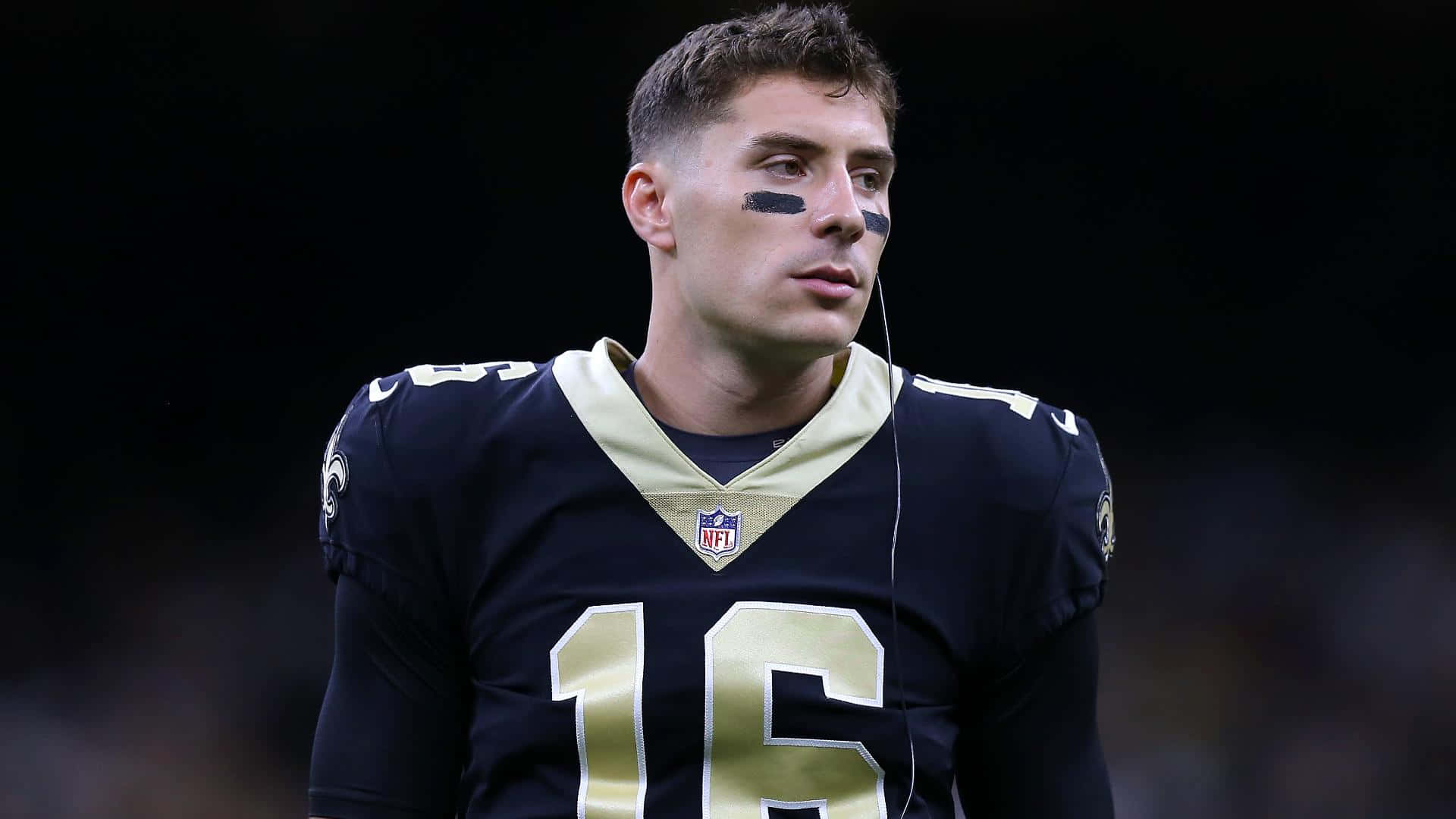  壁紙 — 一位專注的NFL四分衛身穿深藍色和金色16號球衣，在體育場燈光下比賽中凝視前方。