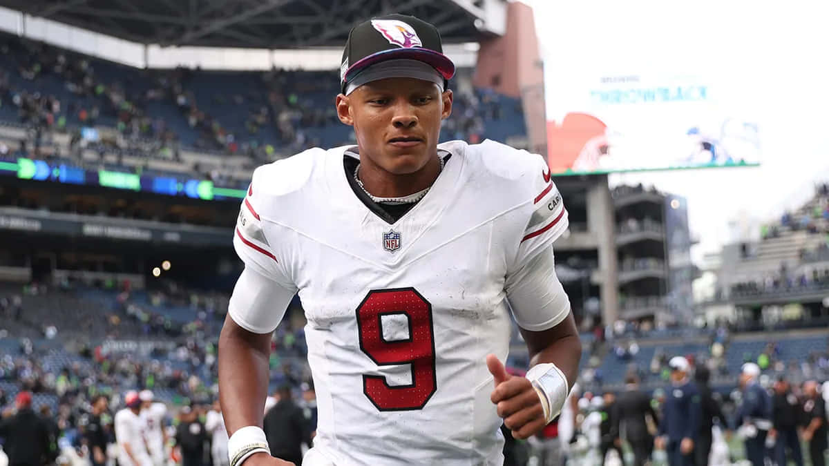  壁紙 — 一位專注的NFL四分衛身穿白色球衣，紅色九號，在球場上奔跑，背景是模糊的觀眾。