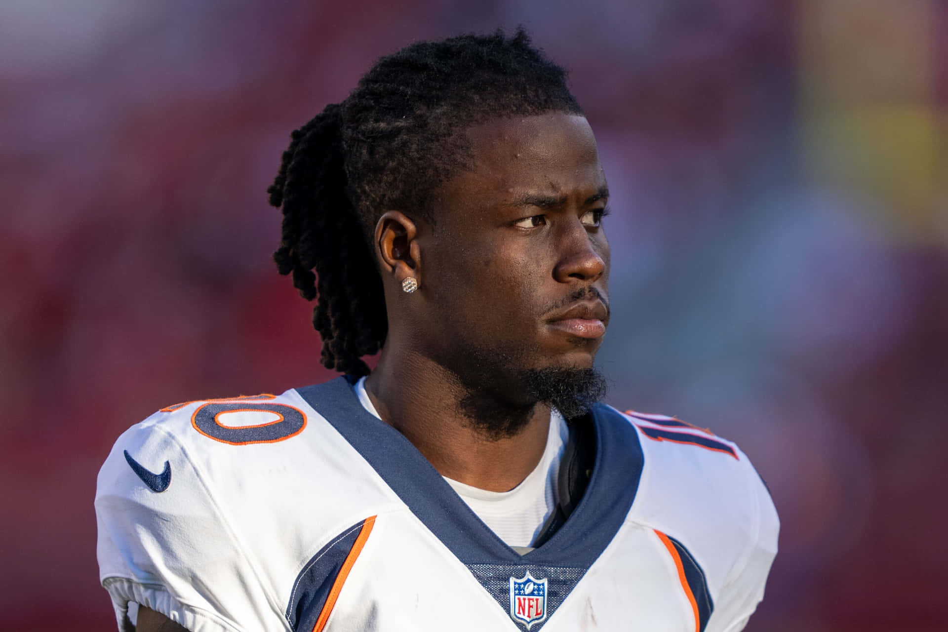  壁紙 — 一名專注的NFL美式足球員身穿白色和橙色球衣，在模糊的體育場背景下凝視鏡頭外。