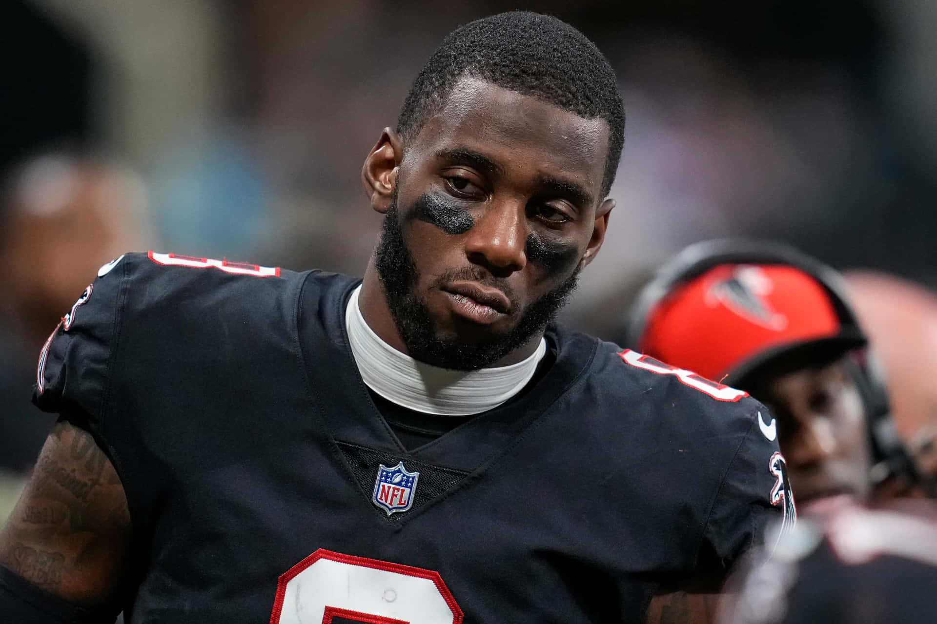  壁紙 — 一位專注的NFL美式足球員，身穿帶有紅色點綴的黑色球衣，並塗有眼部黑色，在比賽中專注地凝視。