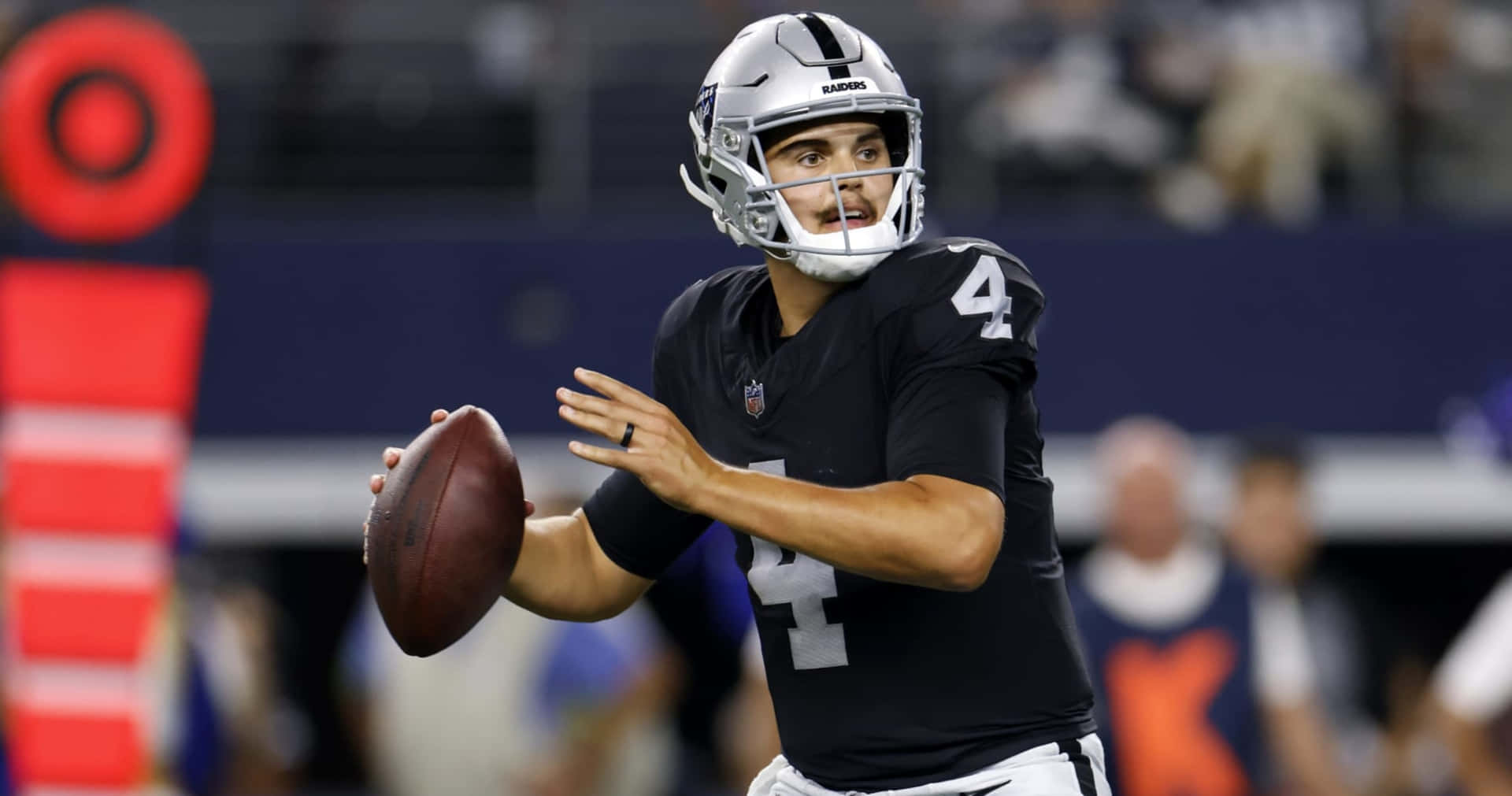 壁紙 — 一位專注的NFL四分衛，身穿黑色和銀色制服，在體育場燈光下準備在比賽中擲出橄欖球。