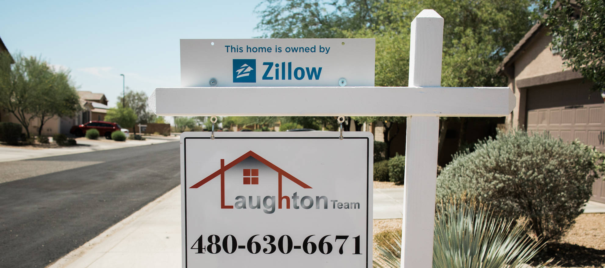 木紋 桌布 壁紙 — Zillow 和 Laughton Team 的白色房地產標誌矗立在陽光明媚的郊區前院，背景有沙漠植物和房屋。