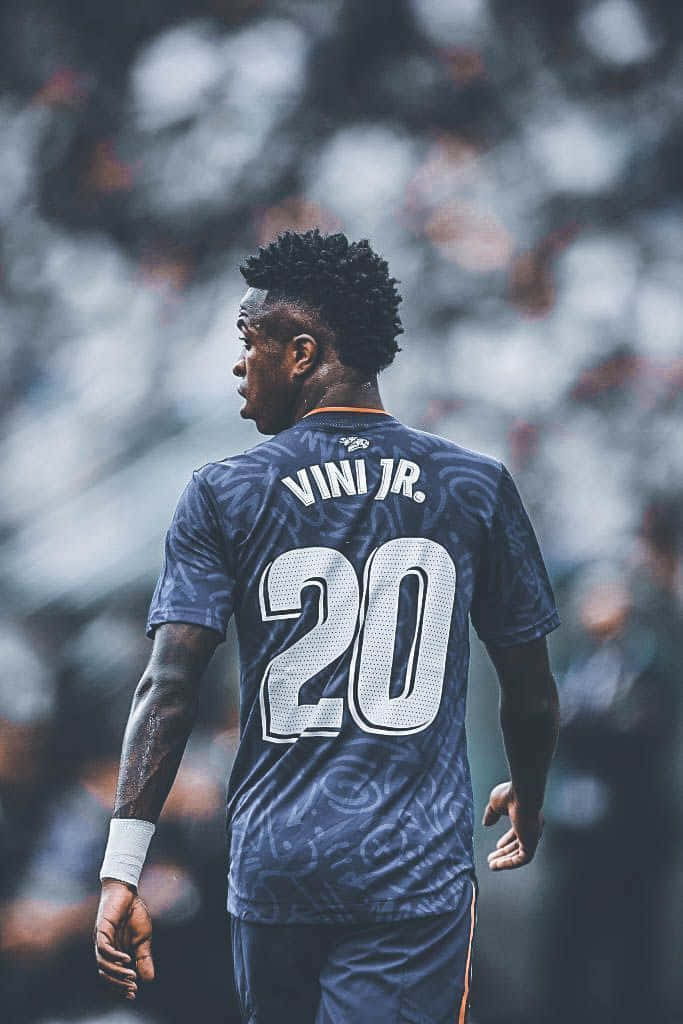 壁紙 — 一名足球員身穿深藍色球衣，背後印有「VINI JR.」和號碼「20」，站在一個觀眾模糊的體育場內。