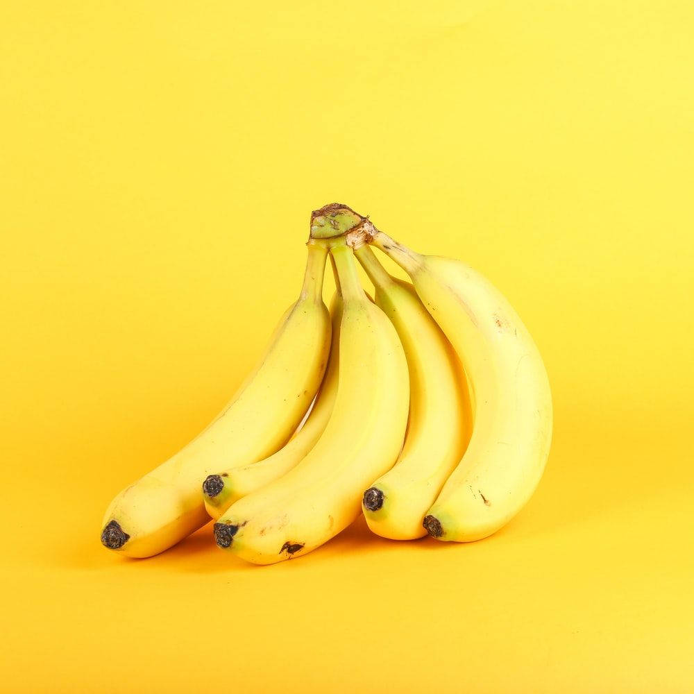 Plantain 桌布 壁紙 — 一串充滿活力的成熟香蕉，帶有輕微的棕色斑點，靠在純黃色背景上。