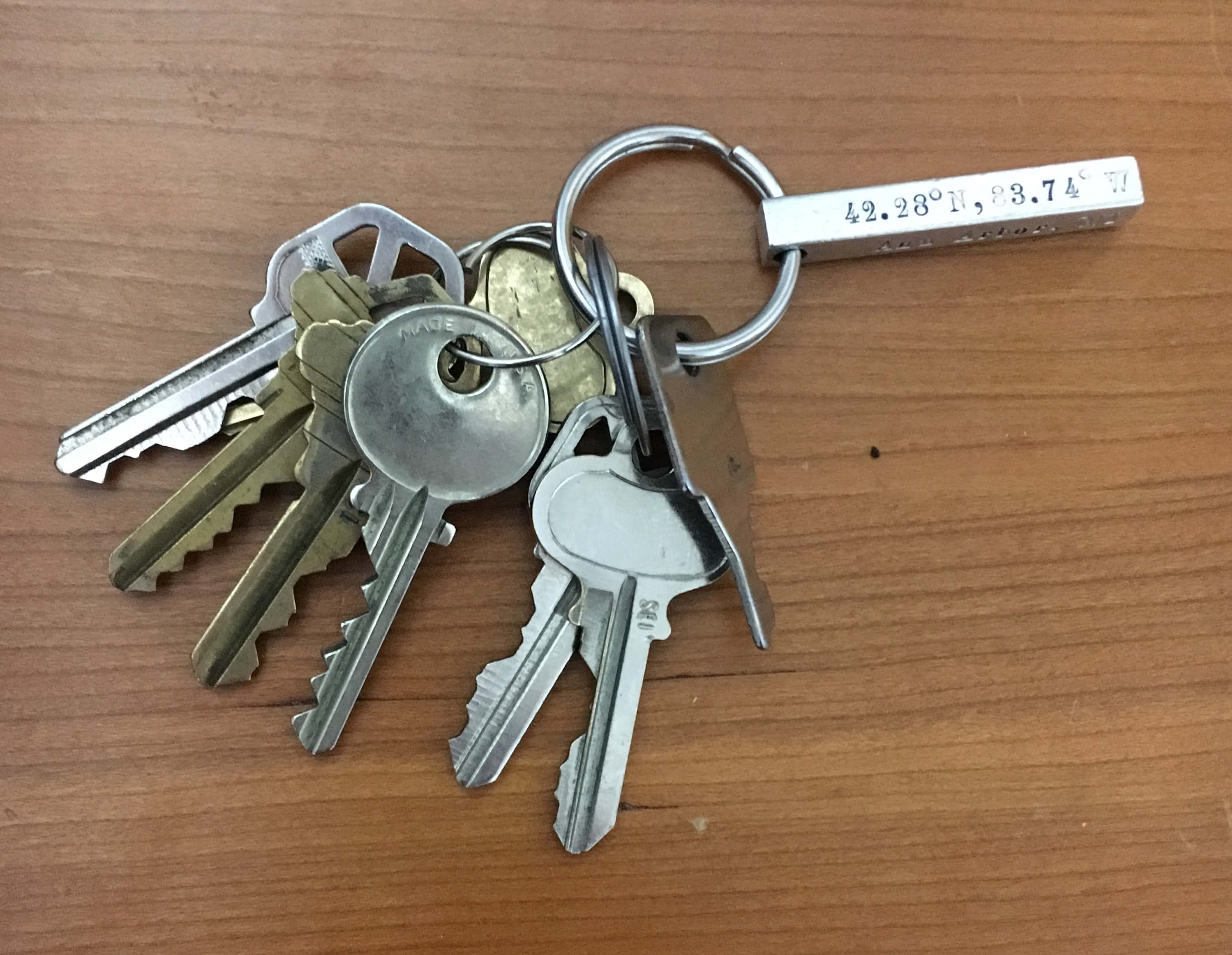 金屬質感 桌布 壁紙 — 一串各式金屬鑰匙掛在一個銀環上，銀環連接到一條刻有地理坐標的棒上，放在溫暖的木質表面上。