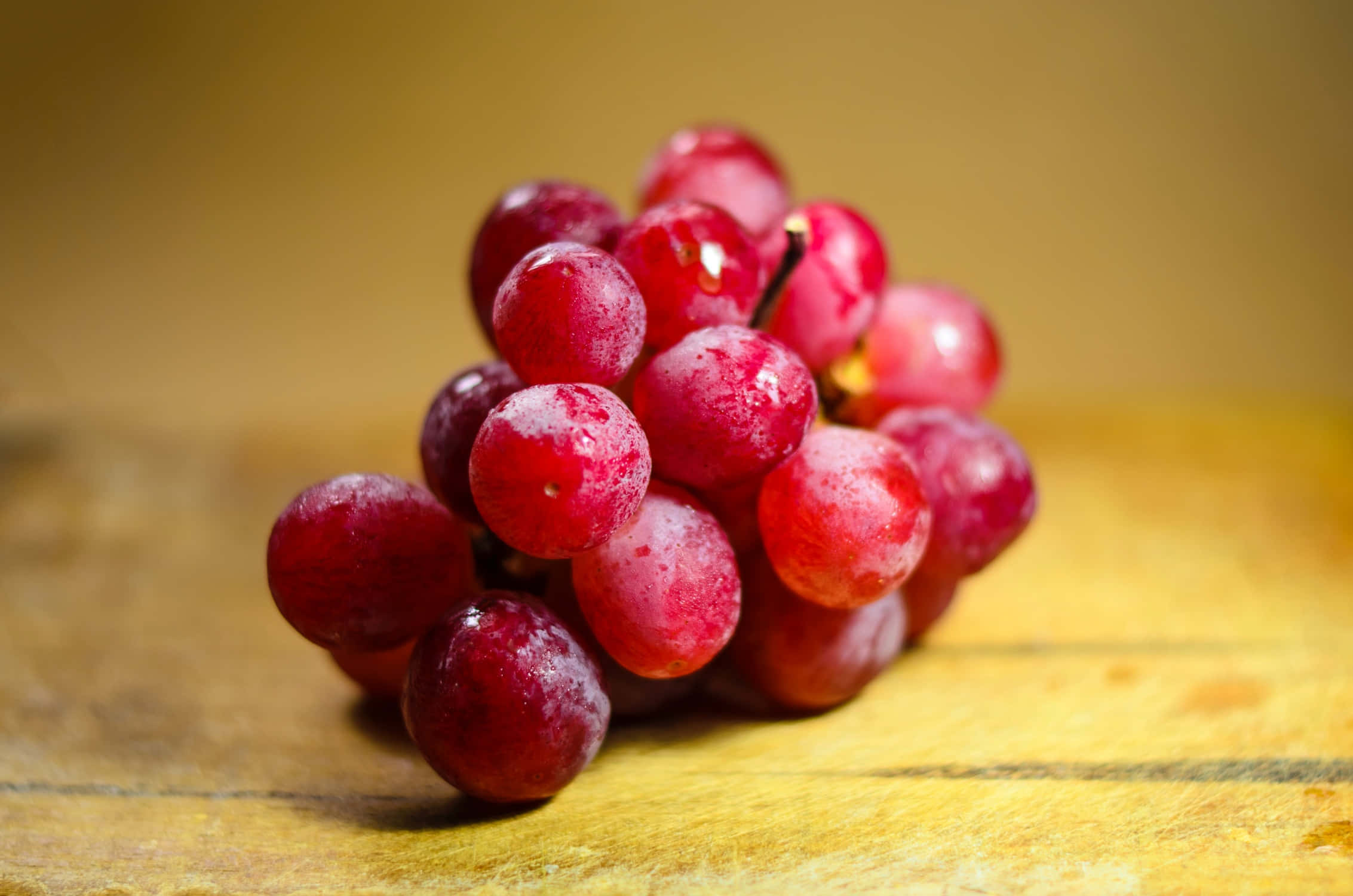 Grape 桌布 壁紙 — 一串飽滿的紅葡萄在柔和的金色光線下，在溫暖的木質表面上閃爍著露珠。