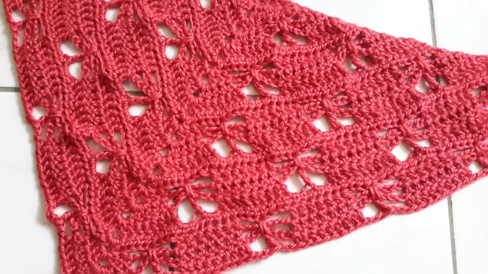 Butterfly Stitch 桌布 壁紙 — 一件充滿活力的珊瑚紅色鉤針編織披肩，帶有複雜的花卉蕾絲圖案，垂落在白色瓷磚地板上。
