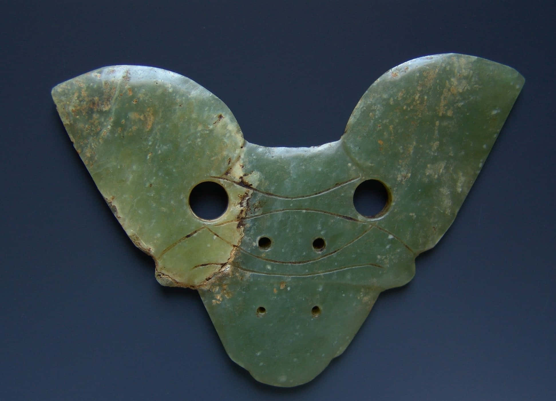 Mystical Artifacts 桌布 壁紙 — 一件古代綠色玉器，形狀像蝴蝶，帶有鑽孔和雕刻線條，背景為深藍色。