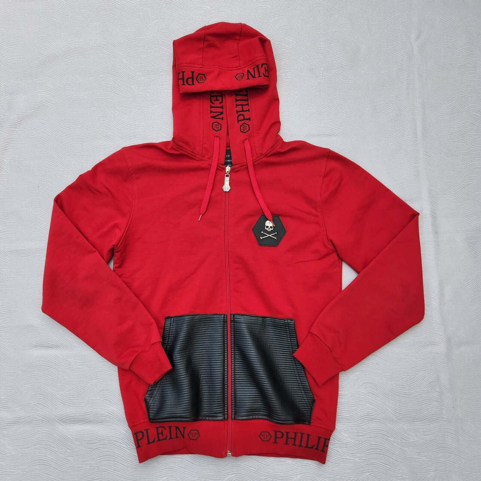 Red Hoodie 桌布 壁紙 — 一件大膽的紅色 Philipp Plein 拉鍊連帽衛衣，帶有黑色皮革拼接和骷髏標誌，平放在有紋理的白色表面上。