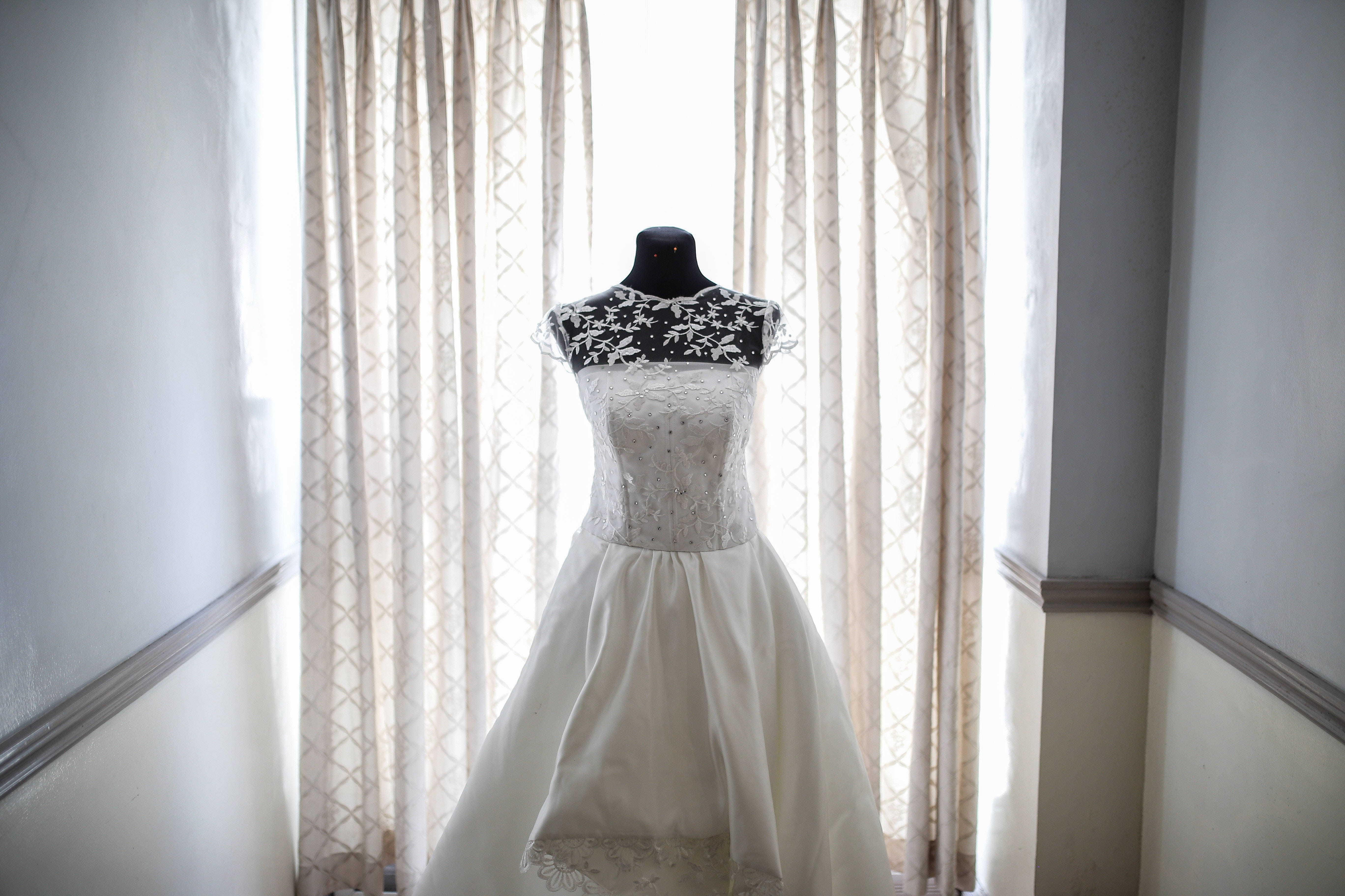 Bridal 桌布 壁紙 — 一件帶有黑色蕾絲細節的白色婚紗掛在一個光線柔和、有圖案窗簾的房間裡的人體模型上。