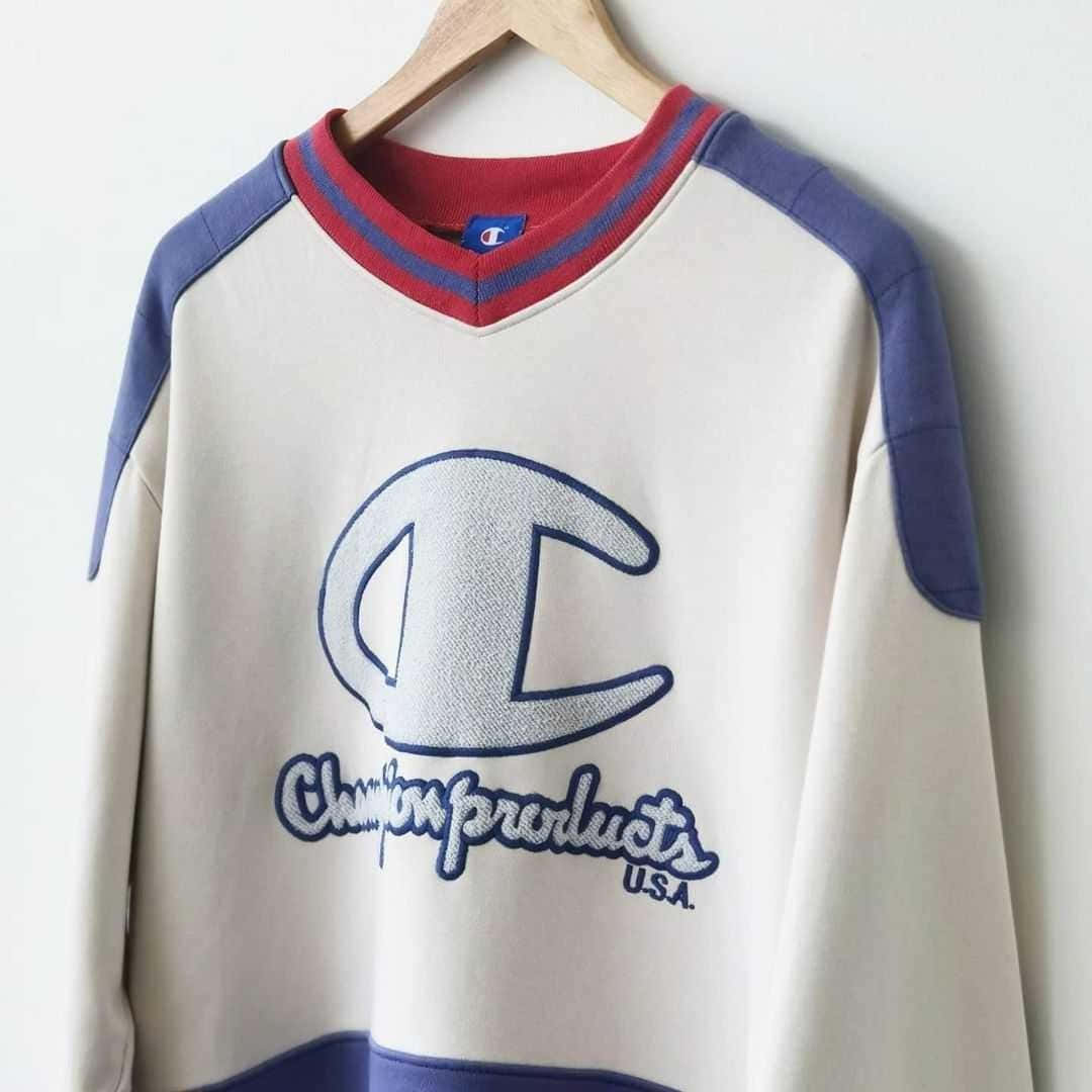 Champion 桌布 壁紙 — 一件復古Champion衛衣，奶油色衣身、藍色袖子、紅色領口，掛在木衣架上，背景是純白牆壁。