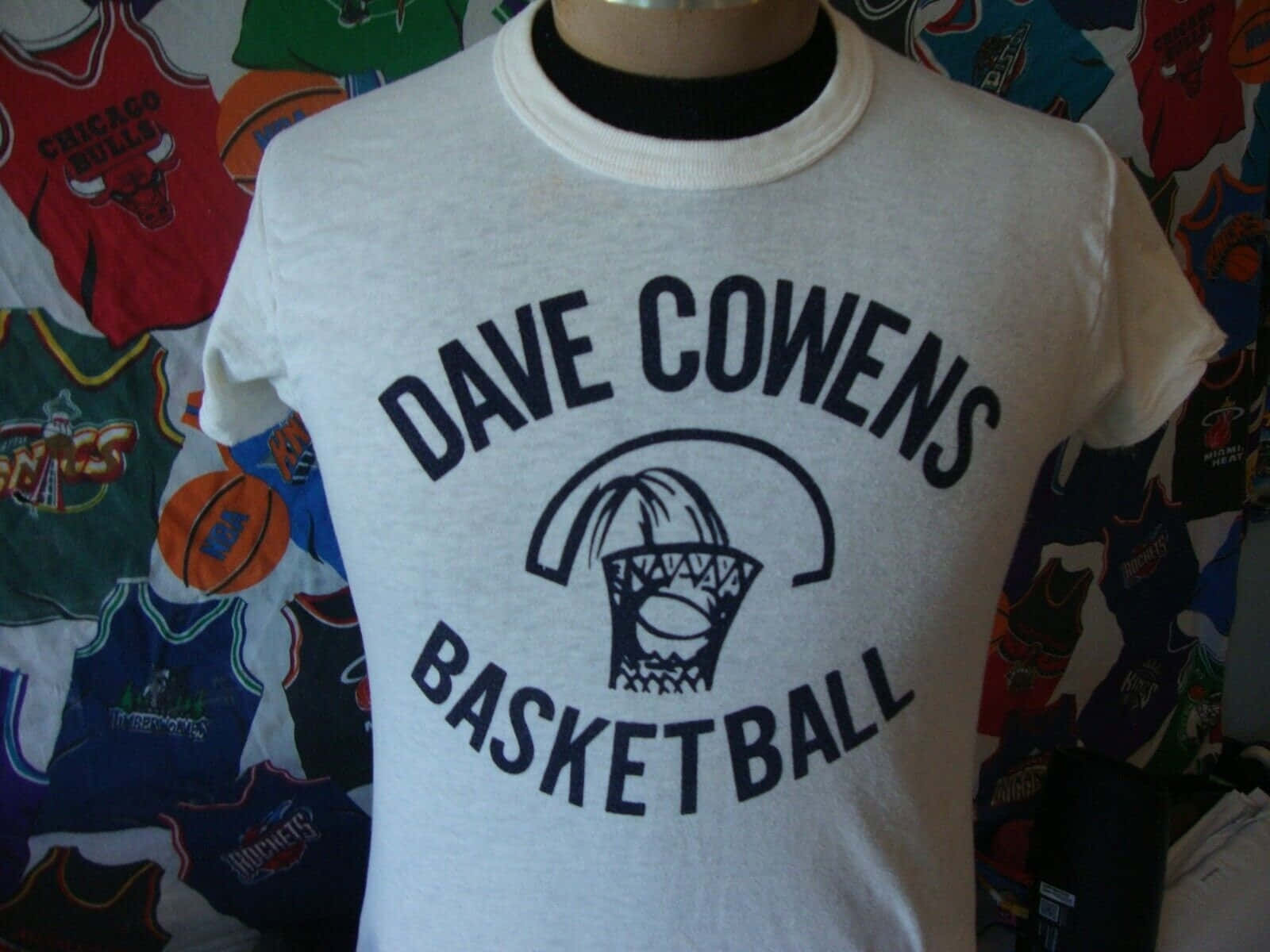 波士頓塞爾提克 桌布 壁紙 — 一件復古白色T恤，上面印有「DAVE COWENS BASKETBALL」和籃框圖案，展示在人體模型上，背景是色彩繽紛的籃球球衣。