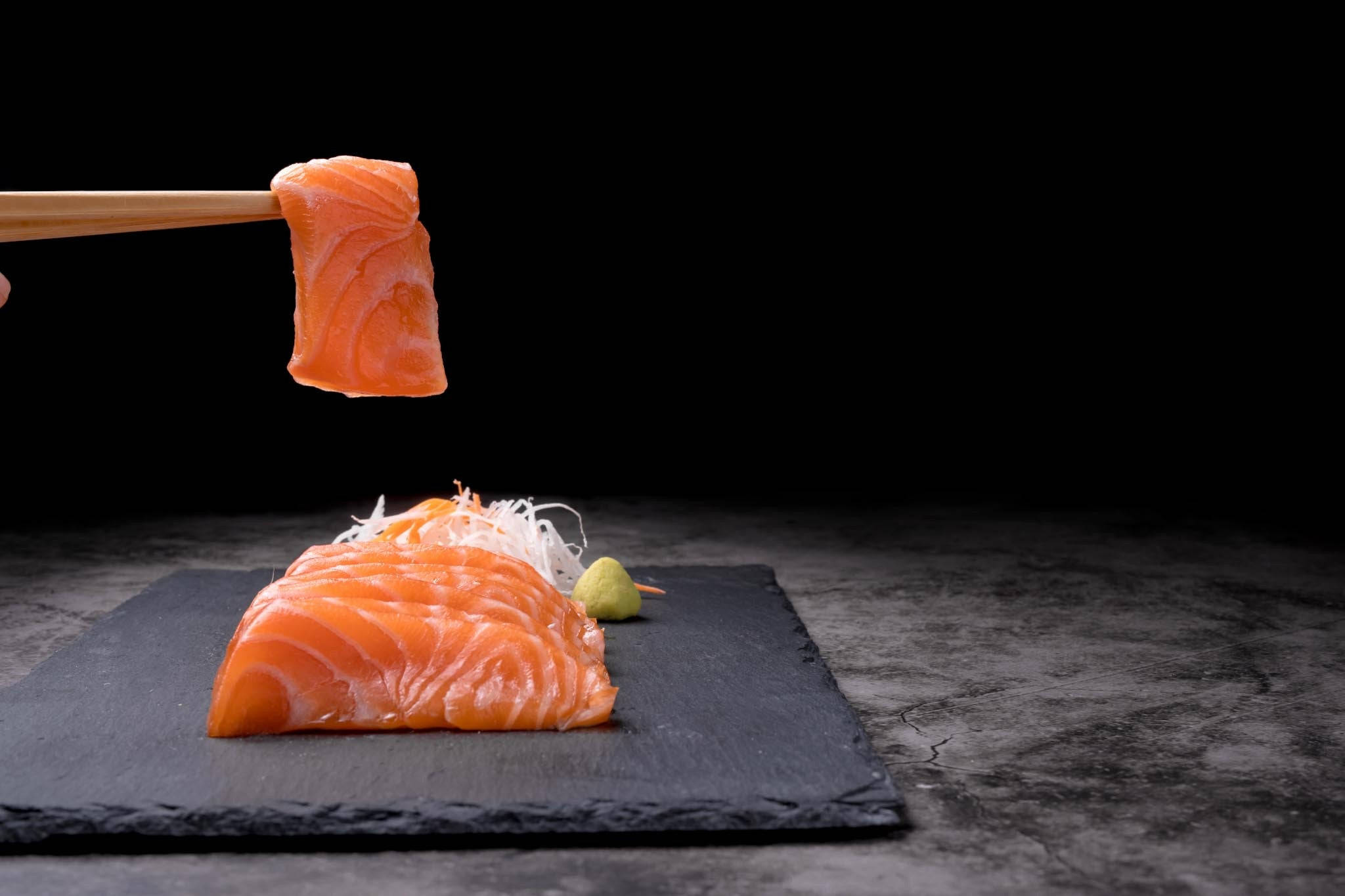 Salmon 桌布 壁紙 — 一件新鮮的鮭魚生魚片被筷子優雅地夾起，放在一個石板盤上，盤上還有更多鮭魚，配以蘿蔔絲和芥末。