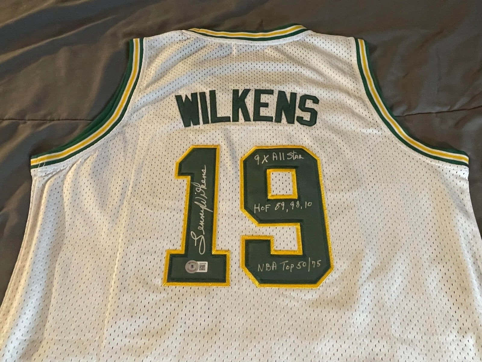 Jersey 桌布 壁紙 — 一件有簽名的白色密爾瓦基公鹿球衣，帶有綠色和金色飾邊，上面印有 'WILKENS' 和號碼 '19'，放在床罩上。