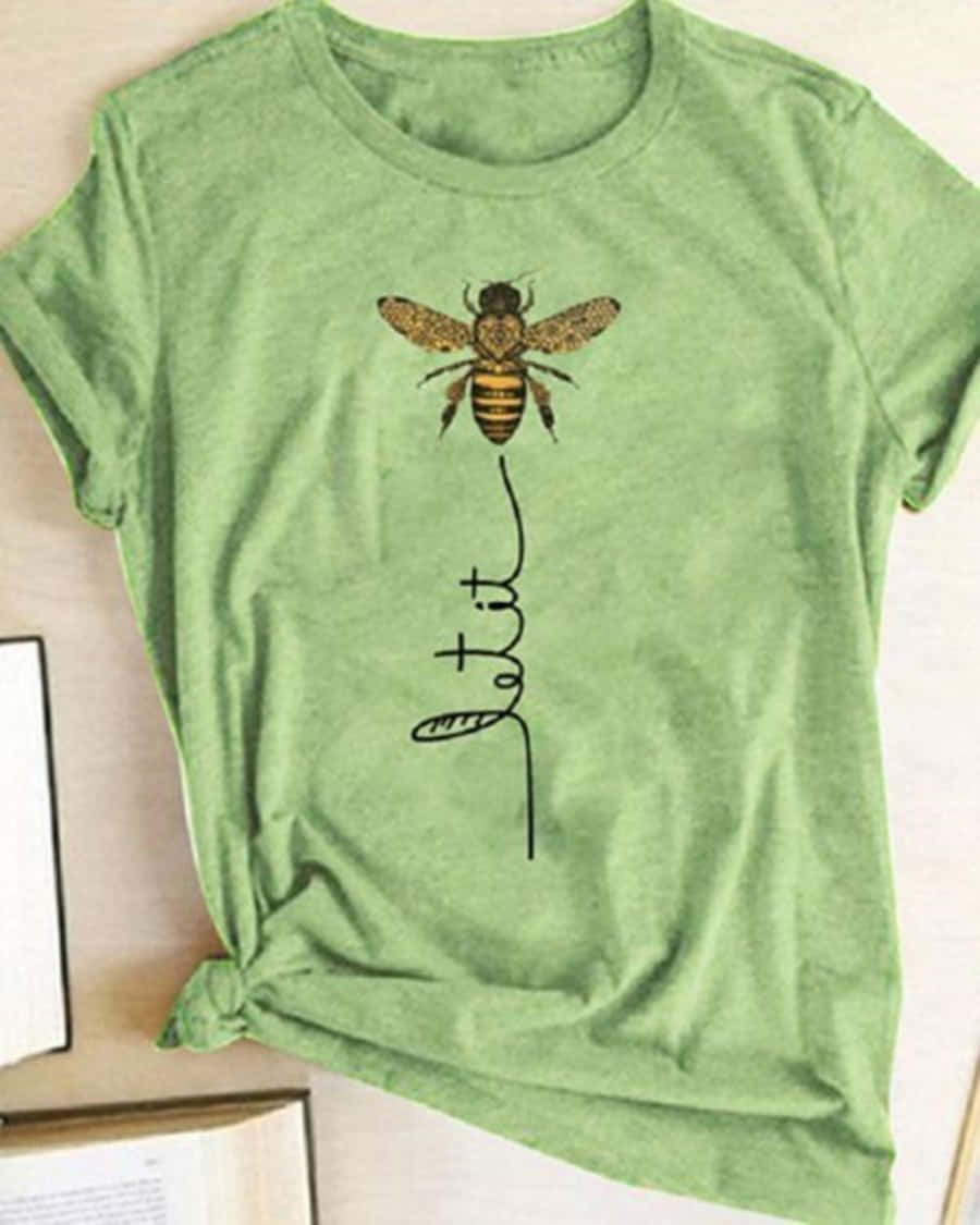 T Shirt 桌布 壁紙 — 一件柔軟的綠色T恤，上面有詳細的蜜蜂插圖和草書短語 let it，隨意地擺放在木質表面上，旁邊是打開的書本。