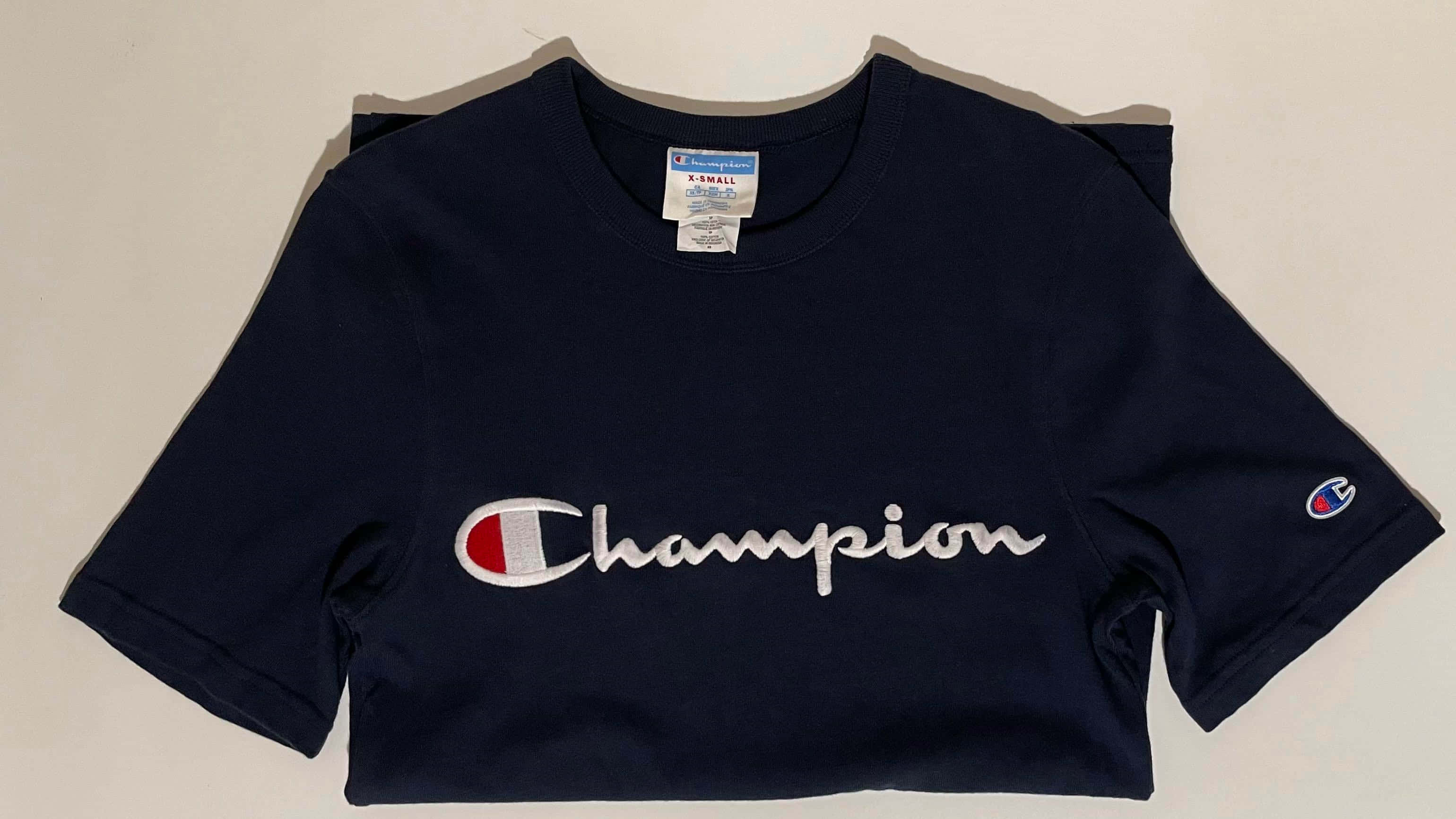 Champion 桌布 壁紙 — 一件海軍藍色Champion短版上衣，帶有白色草書標誌和紅白條紋徽章，平放在淺色背景上。