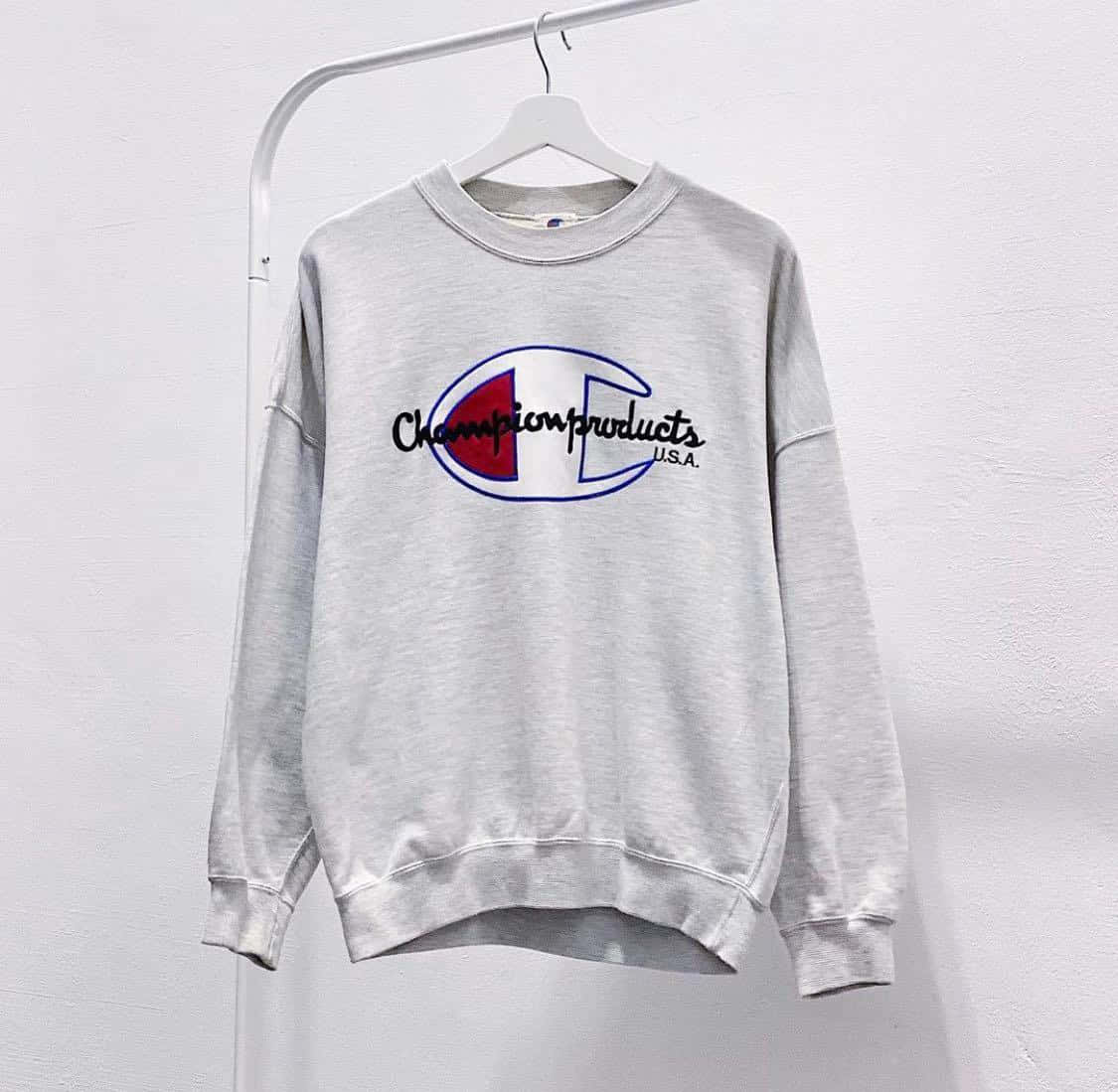 Champion 桌布 壁紙 — 一件淺灰色Champion連帽衛衣，帶有經典標誌，整齊地掛在白色衣架上，背景是素牆。