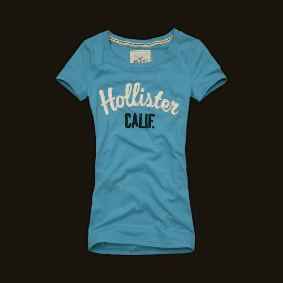 hollister 桌布 壁紙 — 一件淺藍色 Hollister T 恤，上面有白色草書 'Hollister' 和黑色 'CALIF.' 文字，顯示在深棕色背景上。