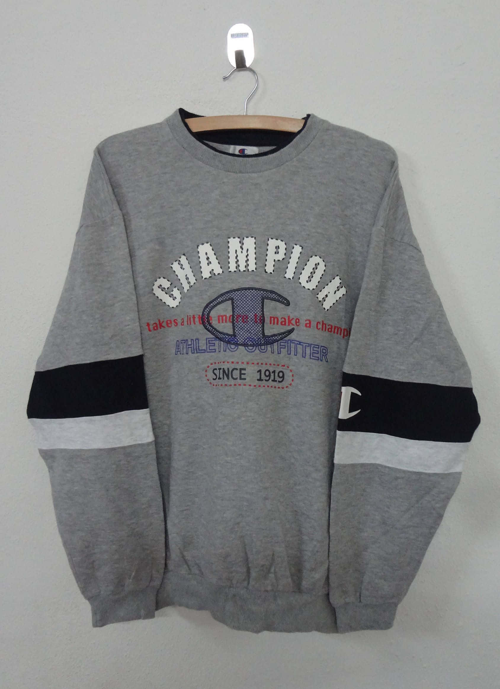 Champion 桌布 壁紙 — 一件灰色Champion連帽衫，帶有黑白條紋袖子，掛在木衣架上，背景是素牆。