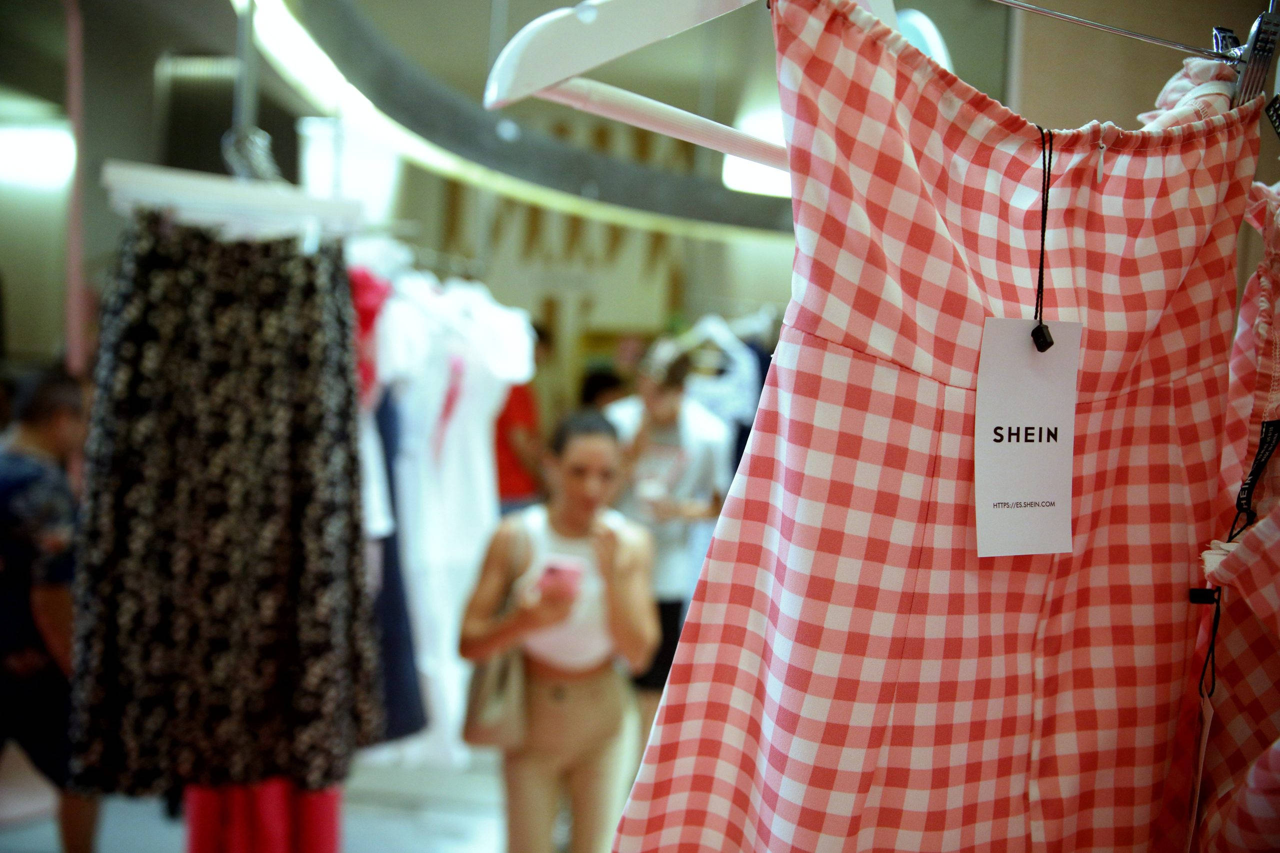 Shein 桌布 壁紙 — 一件粉紅白色格紋連身裙掛著SHEIN標籤，清晰對焦，背景是模糊的服裝店，顧客正在瀏覽。
