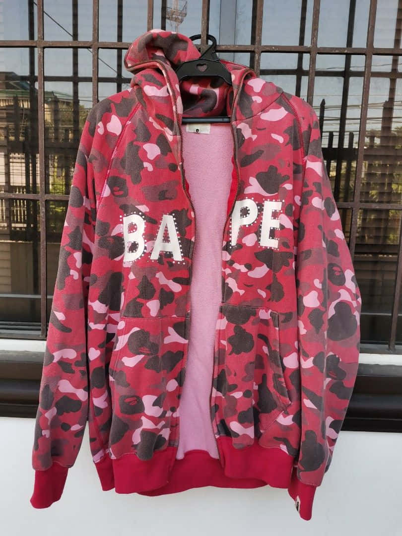 Bape 動態 桌布 壁紙 — 一件紅色、粉紅色和黑色迷彩BAPE連帽上衣掛在衣架上，背景是有城市景觀的鐵窗。