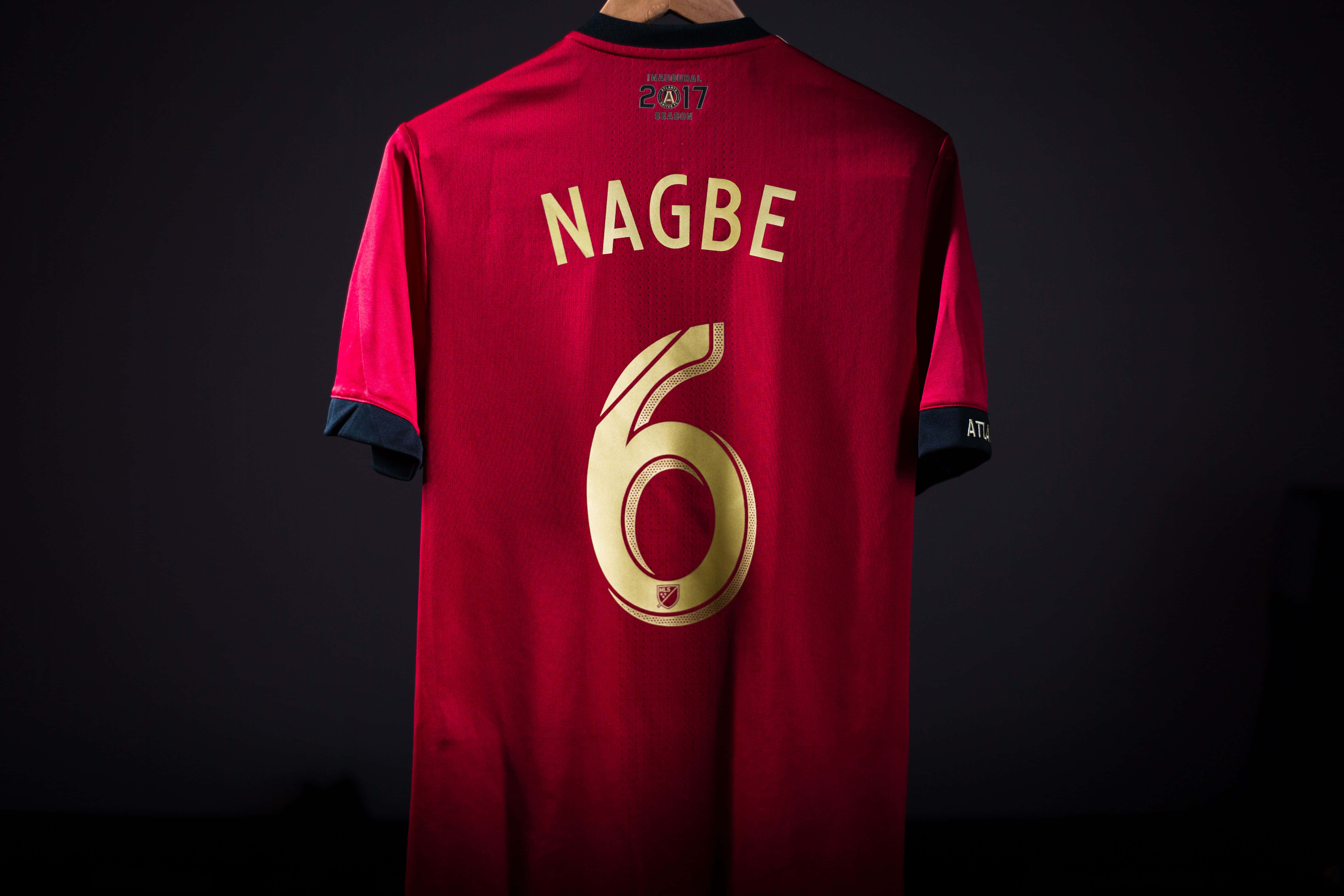 足球 桌布 壁紙 — 一件紅色足球球衣，帶有金色 'NAGBE' 和號碼 '6'，掛在深色背景上。