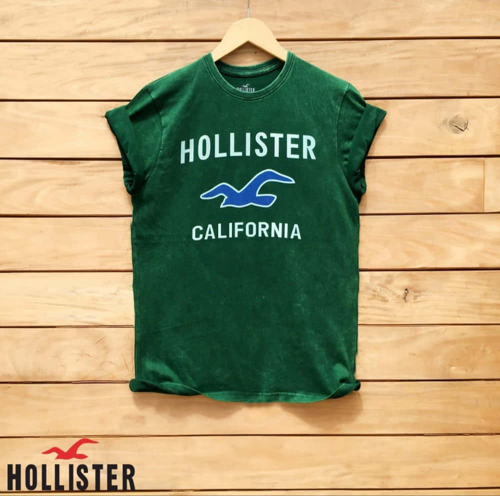 Hollister 桌布 壁紙 — 一件綠色Hollister California T恤，帶有白色文字和藍色海鷗標誌，掛在淺色木條牆上的木衣架上。