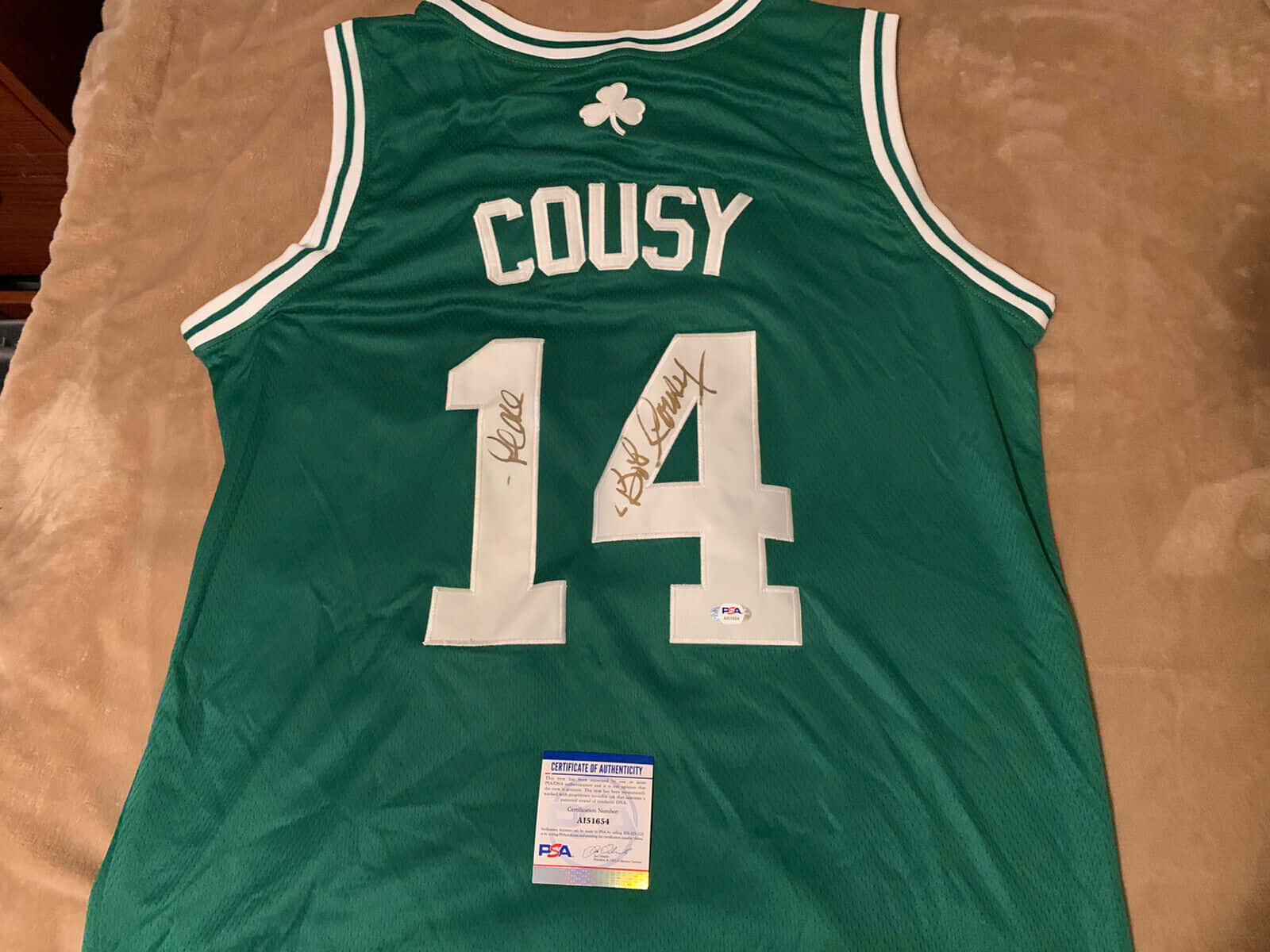 運動 桌布 壁紙 — 一件綠色波士頓塞爾提克球衣，印有白色 'COUSY' 和 14 號，兩次簽名並經 PSA 認證，平放在米色表面上。