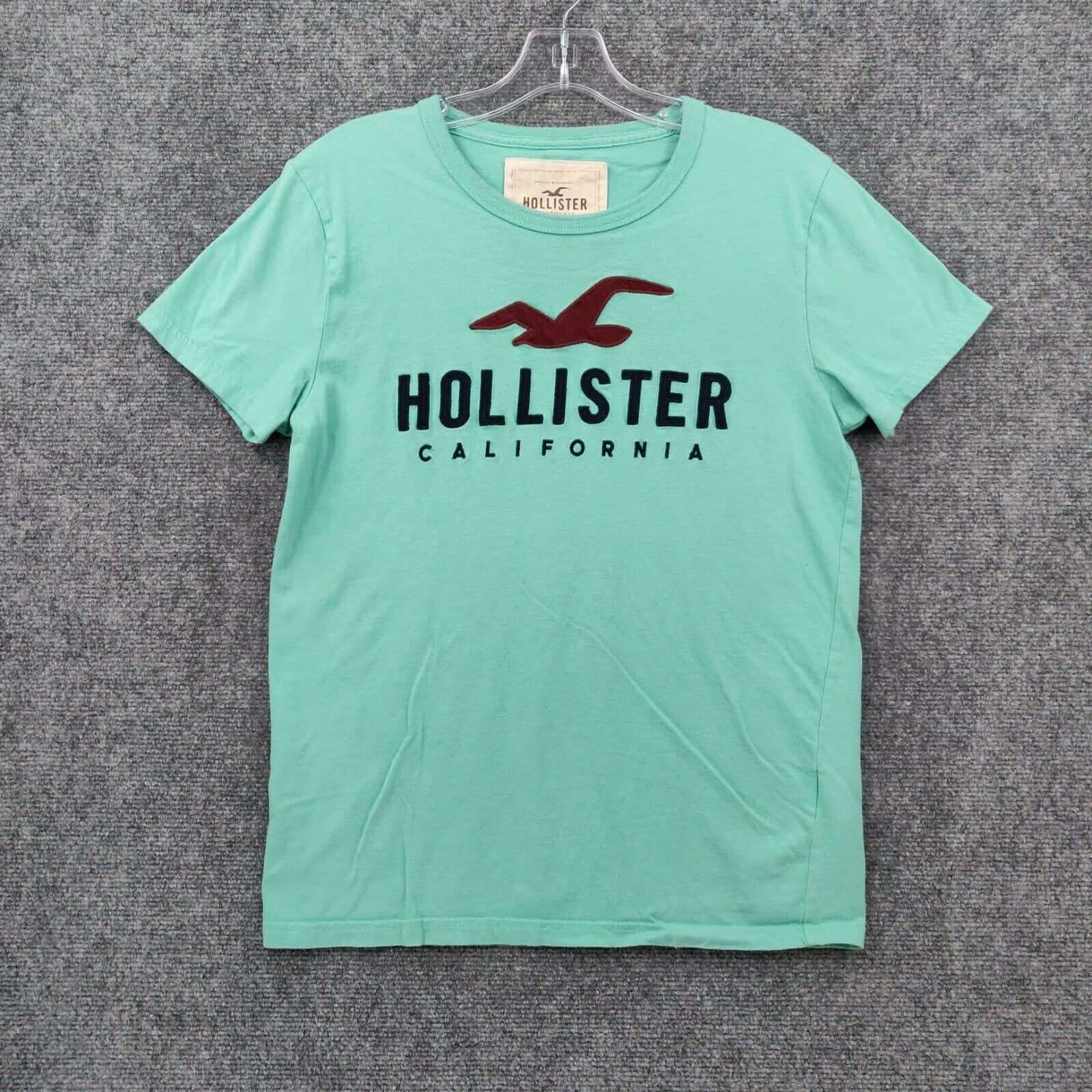 Hollister 桌布 壁紙 — 一件薄荷綠色Hollister T恤，帶有紅色海鷗標誌，掛在透明衣架上，背景為灰色紋理。