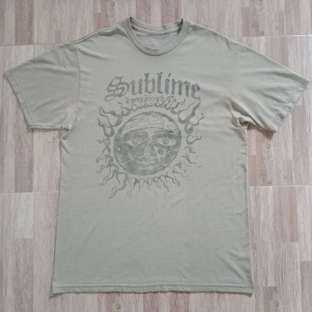 Album Cover 桌布 壁紙 — 一件褪色的橄欖綠色T恤，印有 'Sublime' 樂團圖案，平放在淺色木地板上。