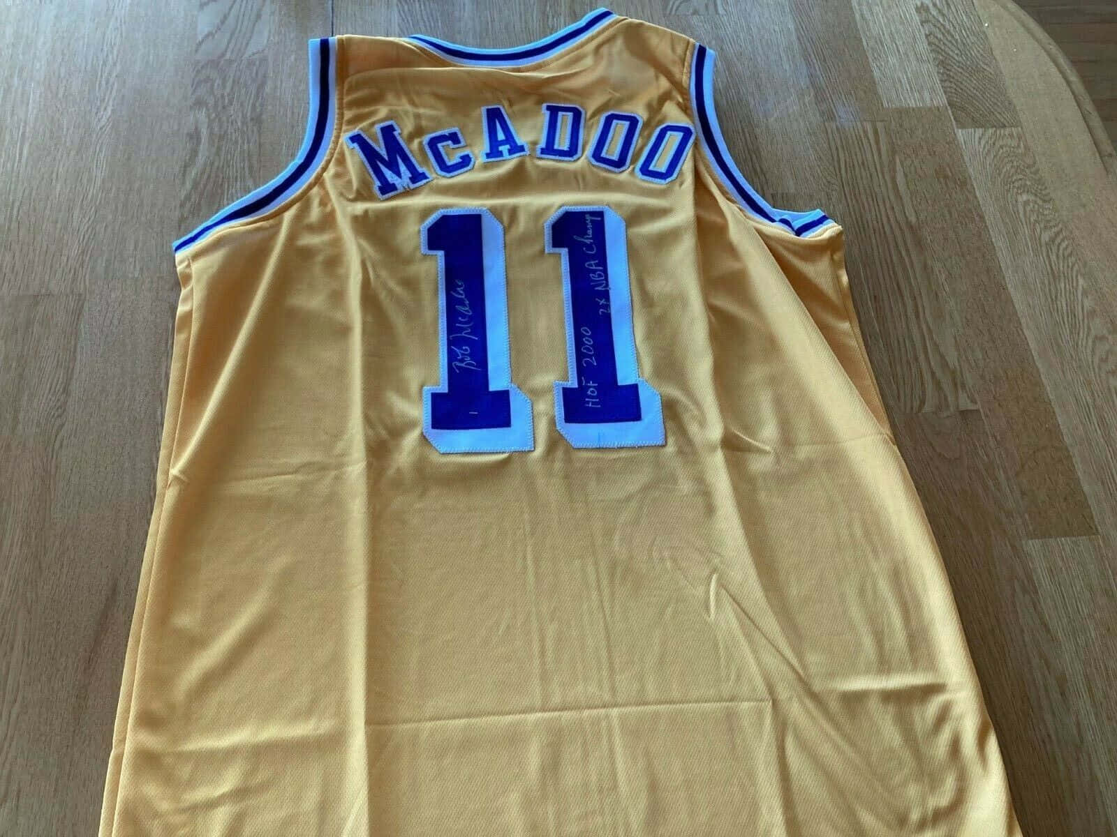 NBA 桌布 壁紙 — 一件金色籃球球衣，上面有藍色 'McADOO' 和號碼 '11'，平放在木地板上，簽名並刻有 'HOF 2000' 和 'NBA Champ'。