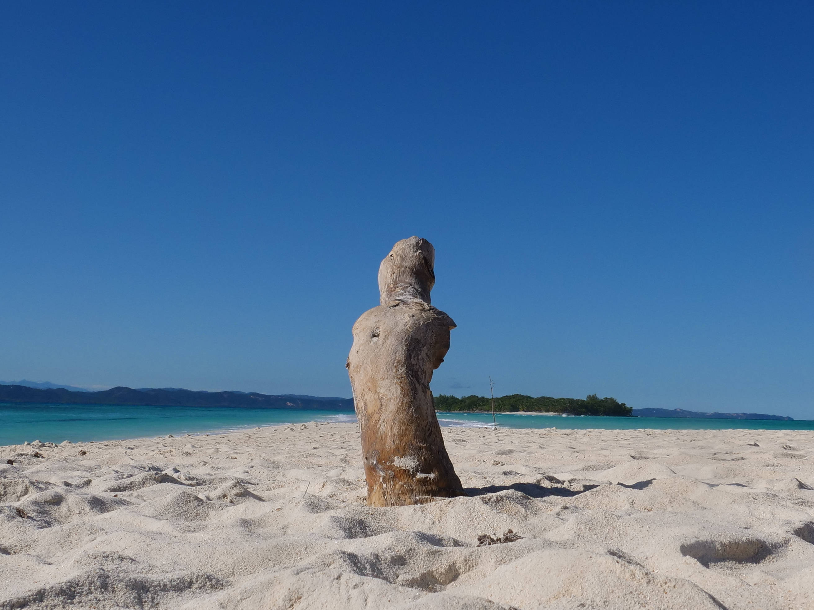 Madagascar 桌布 壁紙 — 一件風化的漂流木雕塑獨自矗立在明亮的白色沙灘上，背景是清澈的藍天、碧綠的海水和遠處的綠色島嶼。