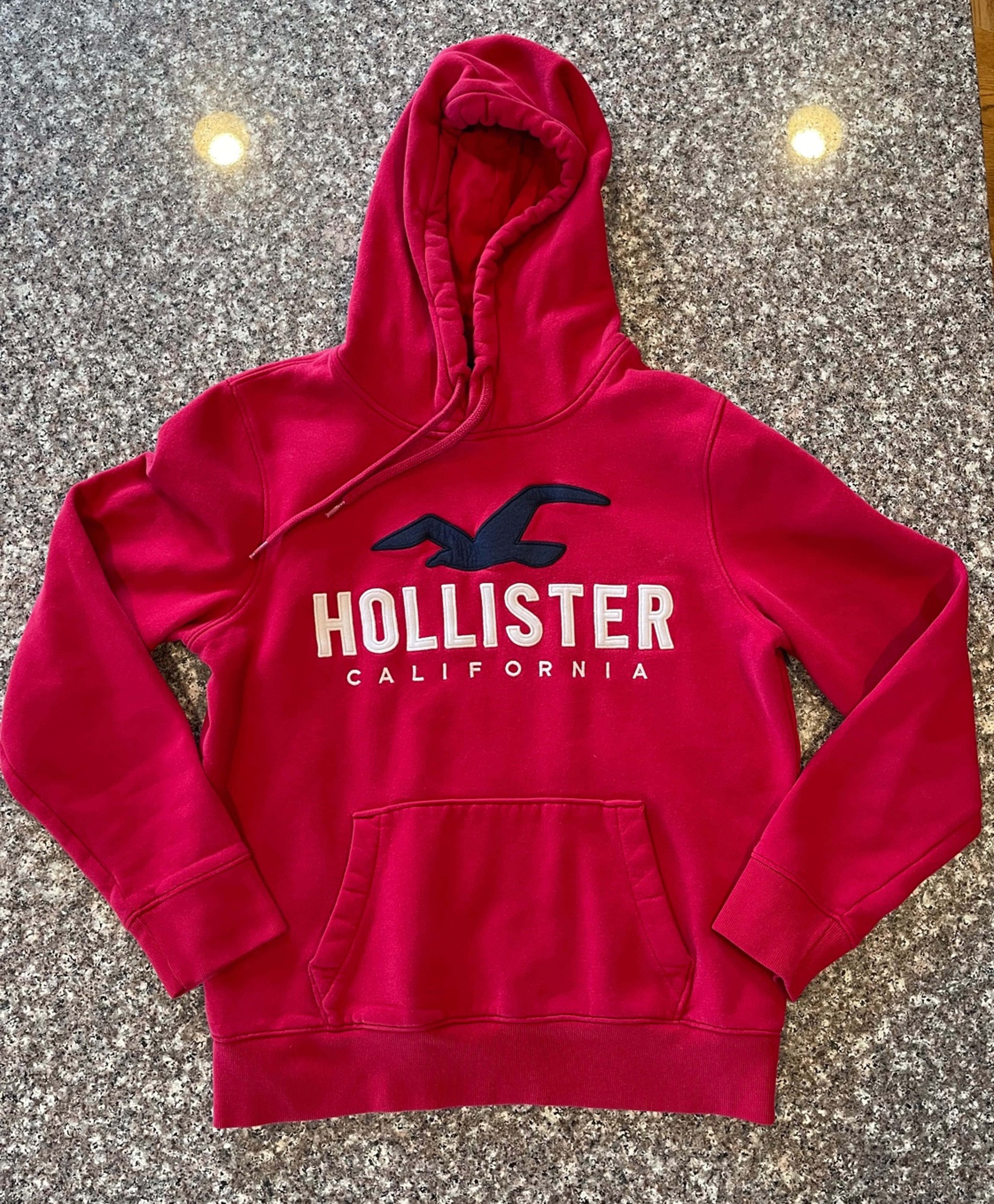 Hollister 桌布 壁紙 — 一件鮮紅色Hollister連帽衫，帶有白色和黑色標誌，平放在有斑點的灰色檯面上，上方是溫暖的吊燈。