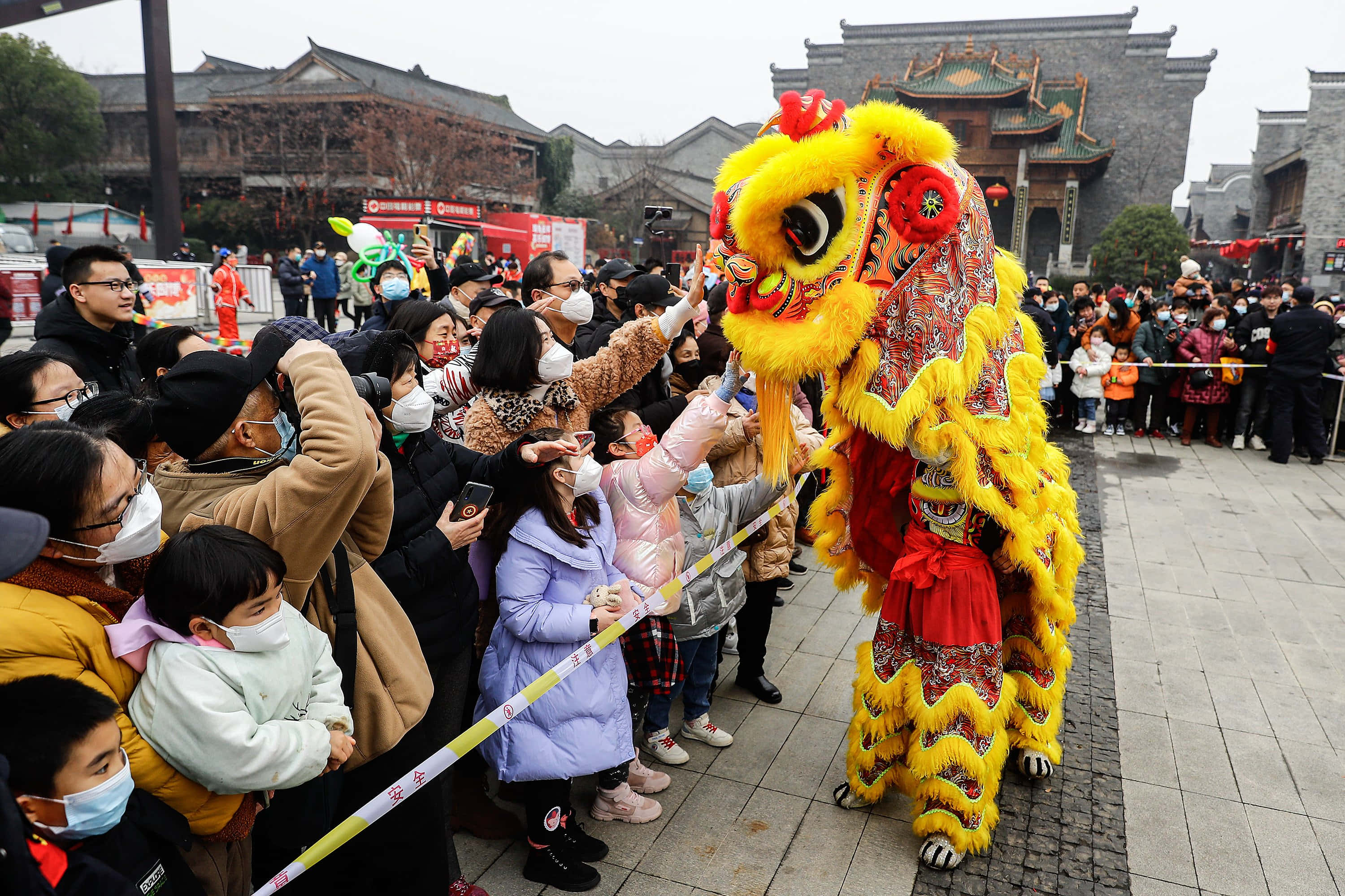 中國 桌布 壁紙 — 一件鮮黃色的舞獅服裝在傳統中式庭院中為一群戴著面具的觀眾表演。