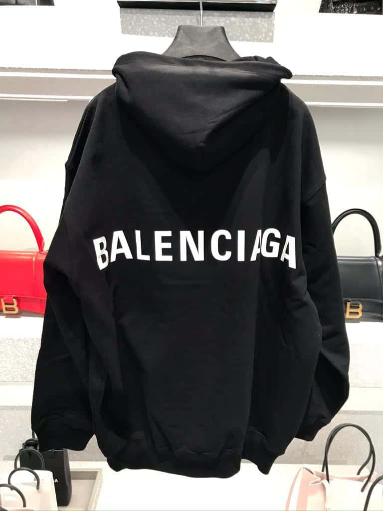 巴黎世家 桌布 壁紙 — 一件黑色Balenciaga連帽上衣，帶有醒目的白色標誌，掛在一個被豪華手提包包圍的人體模型上，呈現時尚的零售展示。