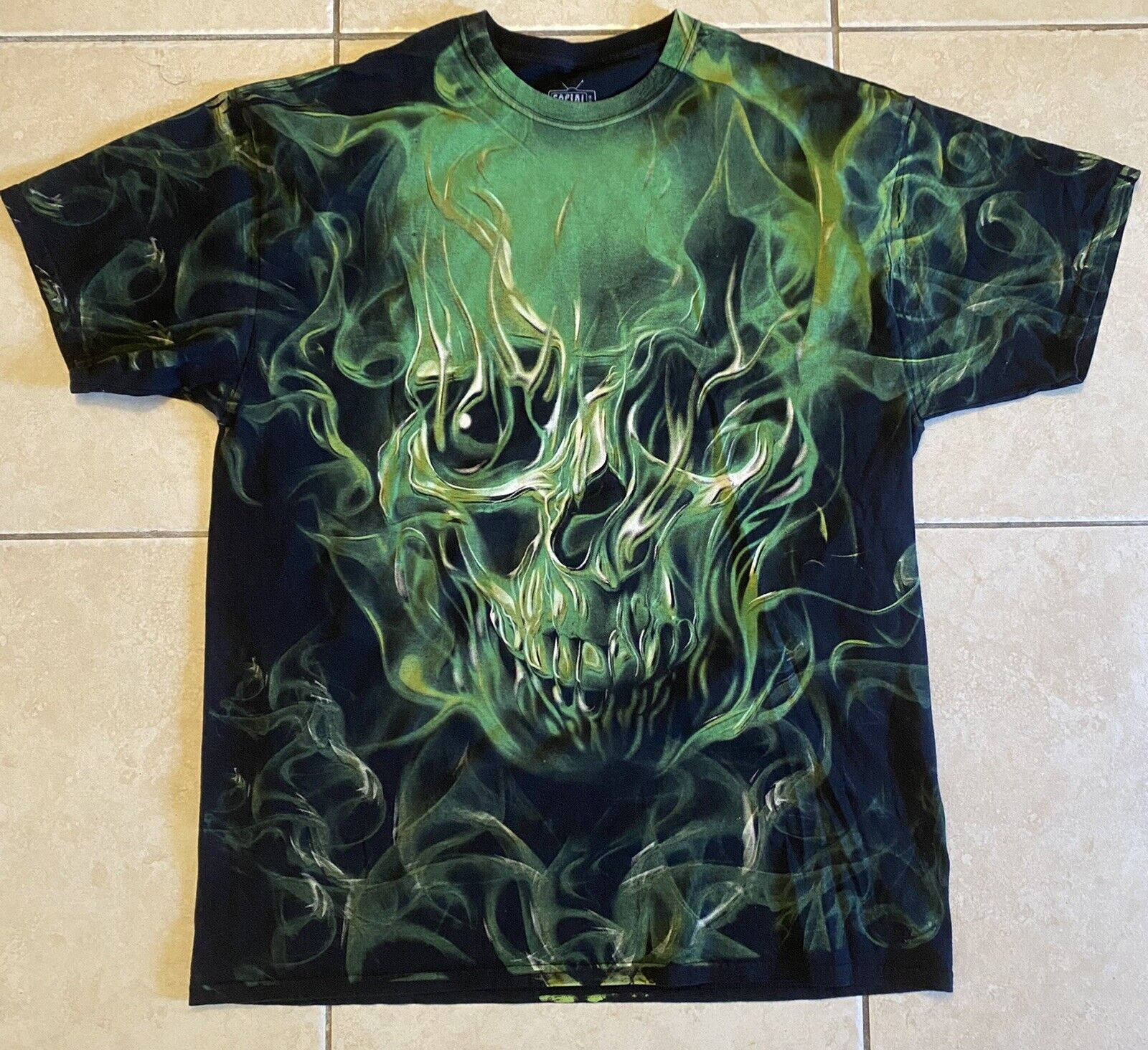 Green Fire Skull 桌布 壁紙 — 一件黑色T恤，上面有鮮綠色火焰骷髏圖案，平放在米色瓷磚地板上。