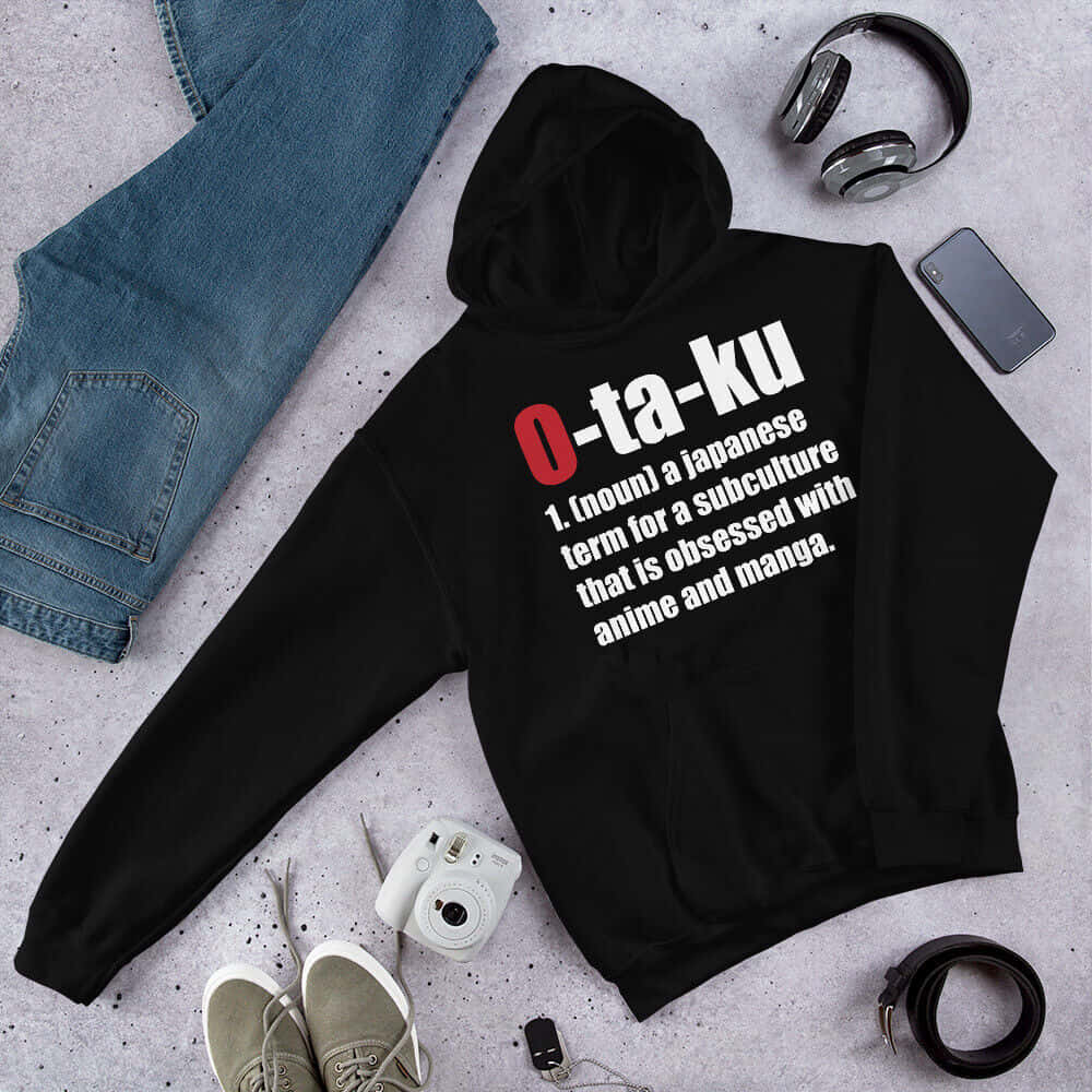 Otaku 桌布 壁紙 — 一件黑色連帽上衣，上面用紅色和白色印有「O-ta-ku」的定義，平放在斑駁的灰色表面上，周圍環繞著牛仔褲、運動鞋、耳機和相機。