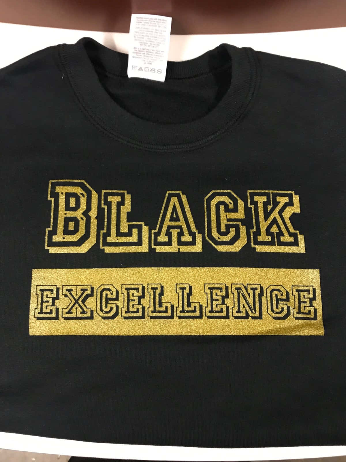 Black Excellence 桌布 壁紙 — 一件黑色連帽衫，上面有粗體金色閃粉文字寫著 BLACK EXCELLENCE，展示在平面上。