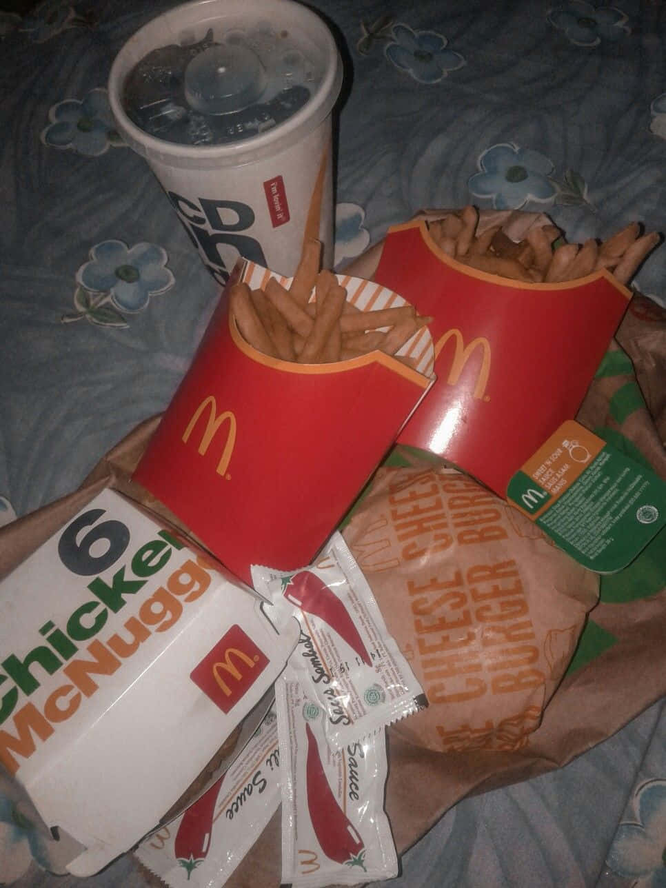 Chicken Nuggets 桌布 壁紙 — 一份凌亂的麥當勞餐點，包括麥克雞塊、薯條和飲料，放在一張花卉圖案的床罩上。