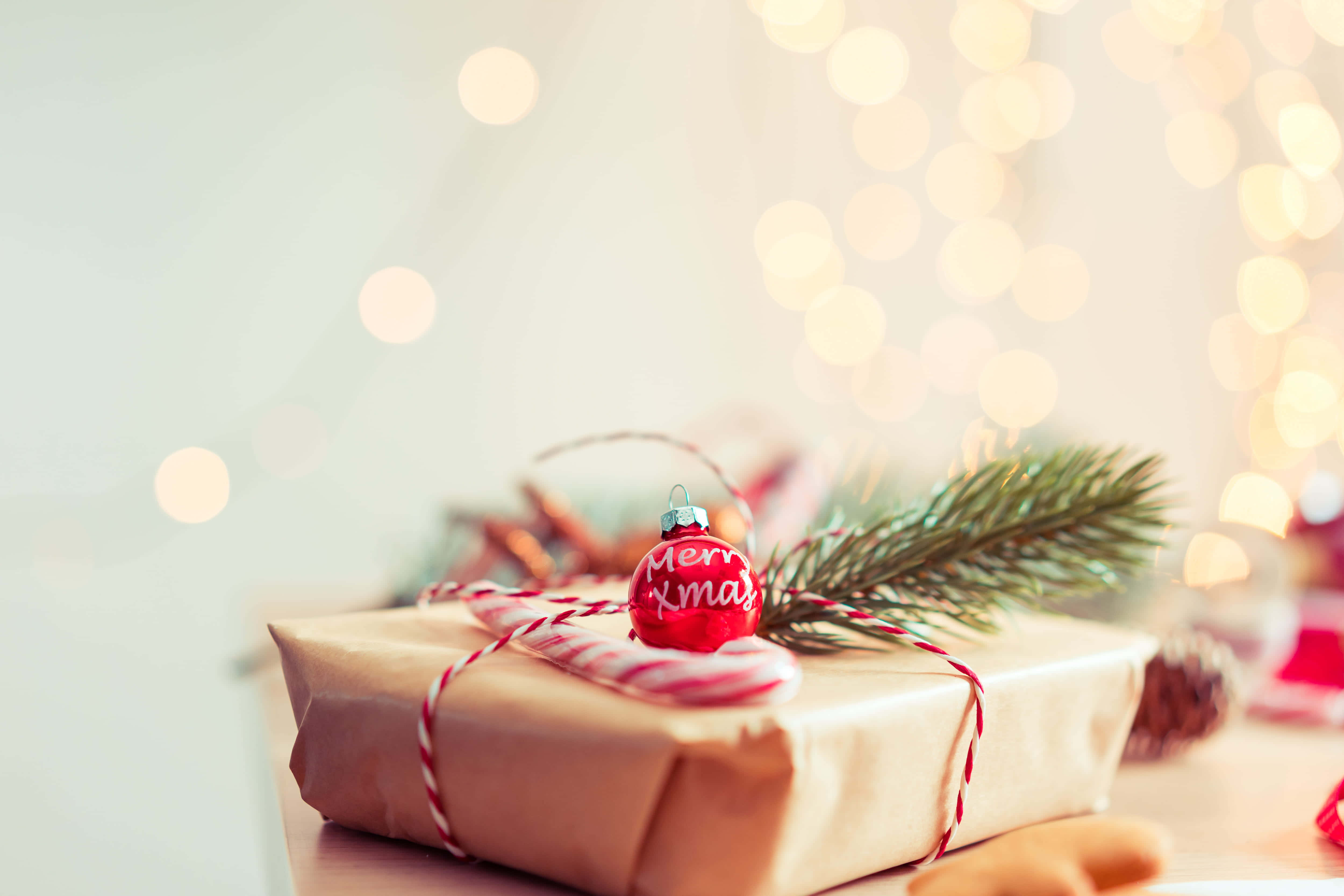 Christmas Gift 桌布 壁紙 — 一份精美包裝的聖誕禮物，飾有紅色裝飾品、拐杖糖和松樹枝，背景是柔和閃爍的燈光。