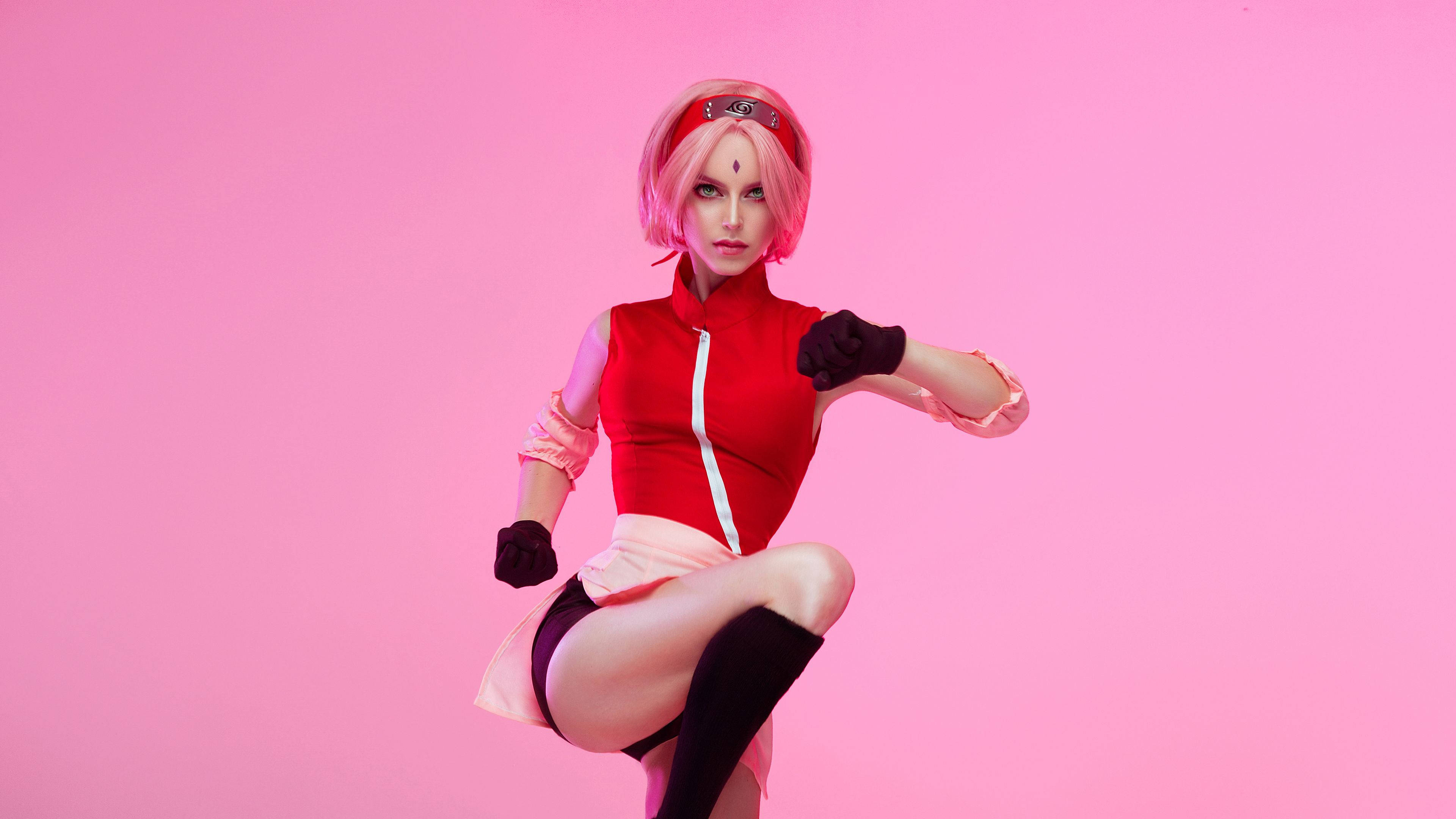 Sakura Haruno 桌布 壁紙 — 一位Cosplayer穿著紅色和粉紅色的火影忍者服裝，在純粉紅色背景前擺出動態武術姿勢。
