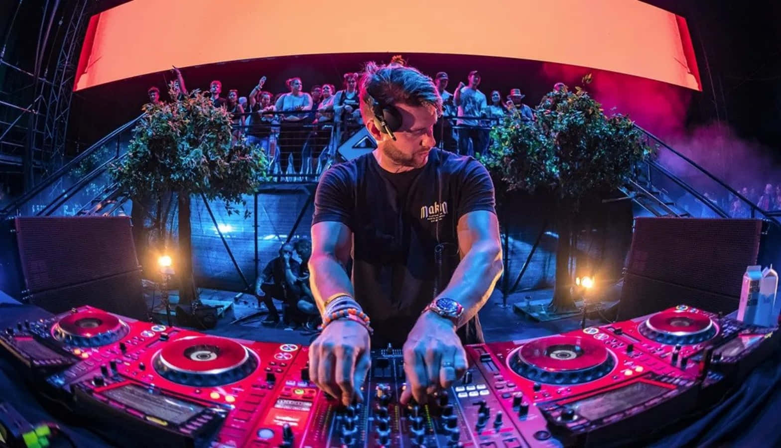 DJ 桌布 壁紙 — 一位DJ在充滿活力的夜店中，在戲劇性的燈光下，用發光的紅色設備和身後的人群激烈地混合音樂。