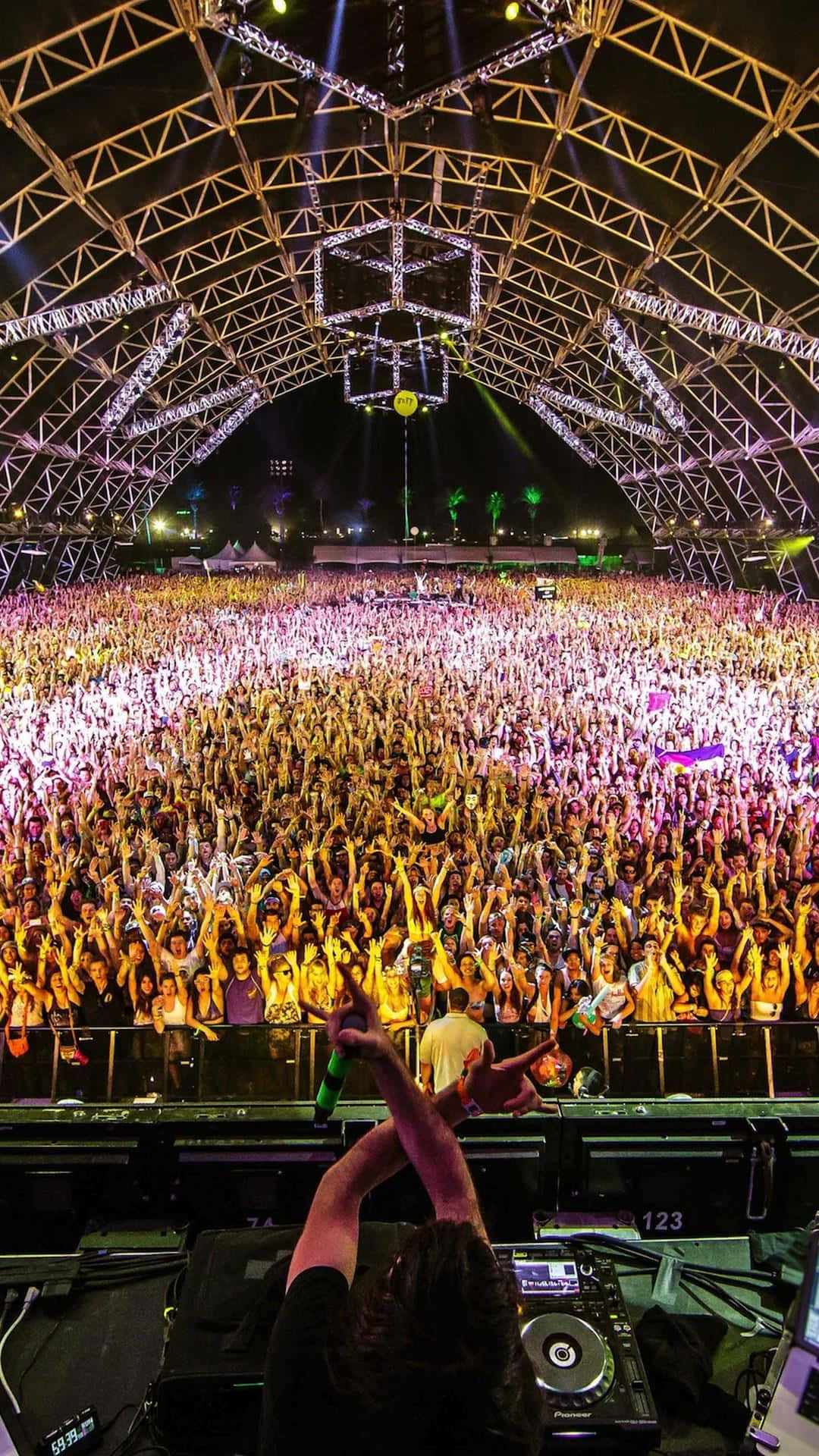 Coachella 桌布 壁紙 — 一位DJ在夜間俯瞰著發光桁架結構下歡呼的龐大音樂會人群。