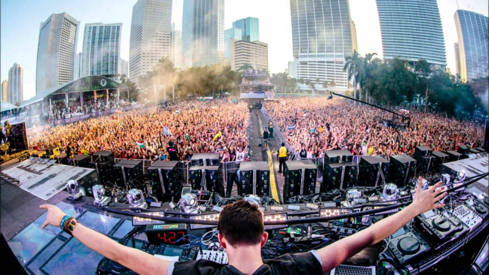 Tomorrowland 桌布 壁紙 — 一位DJ張開雙臂，在摩天大樓下的戶外音樂節指揮著龐大的人群。